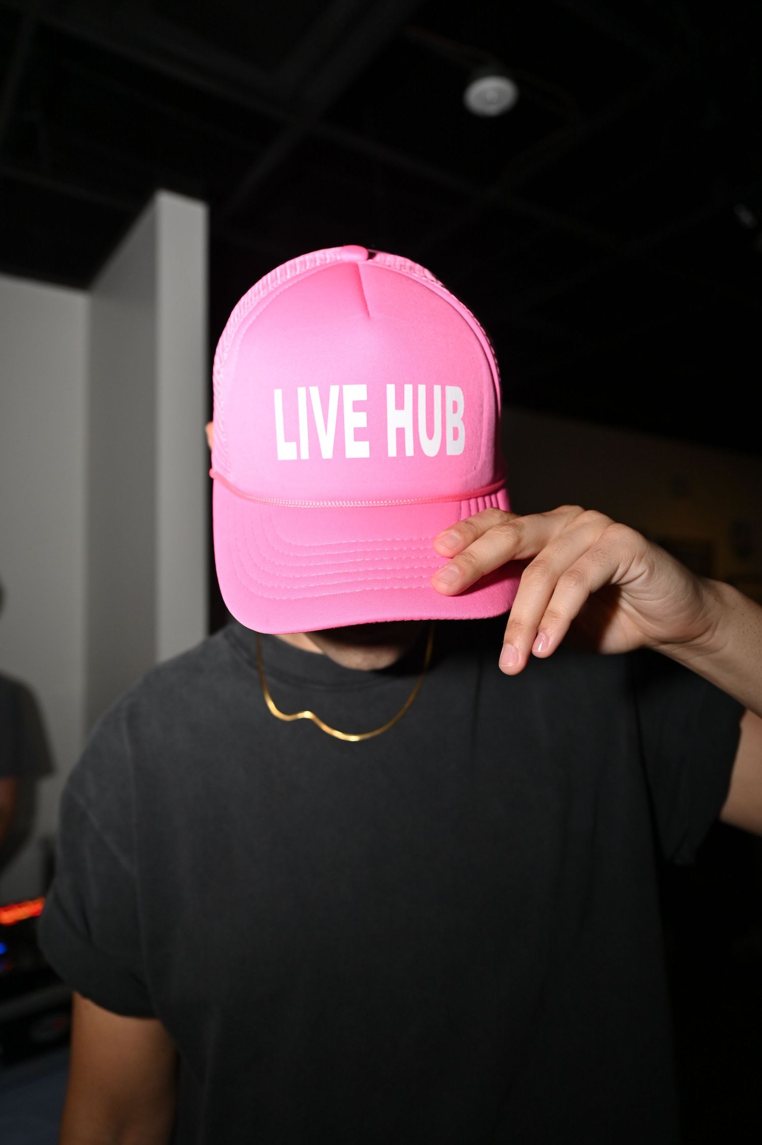 Person wearing a pink Live Hub trucker hat, black t-shirt, and gold chain necklace indoors.