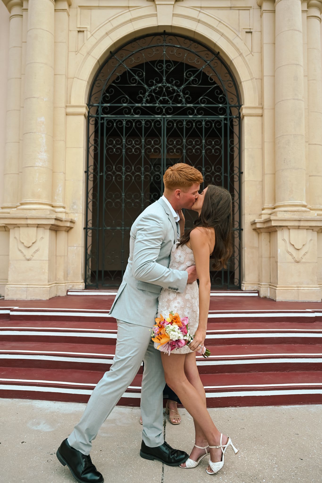 Newlywed couple sharing a romantic kiss on church steps, groom in gray suit, bride in white dress holding colorful bouquet.