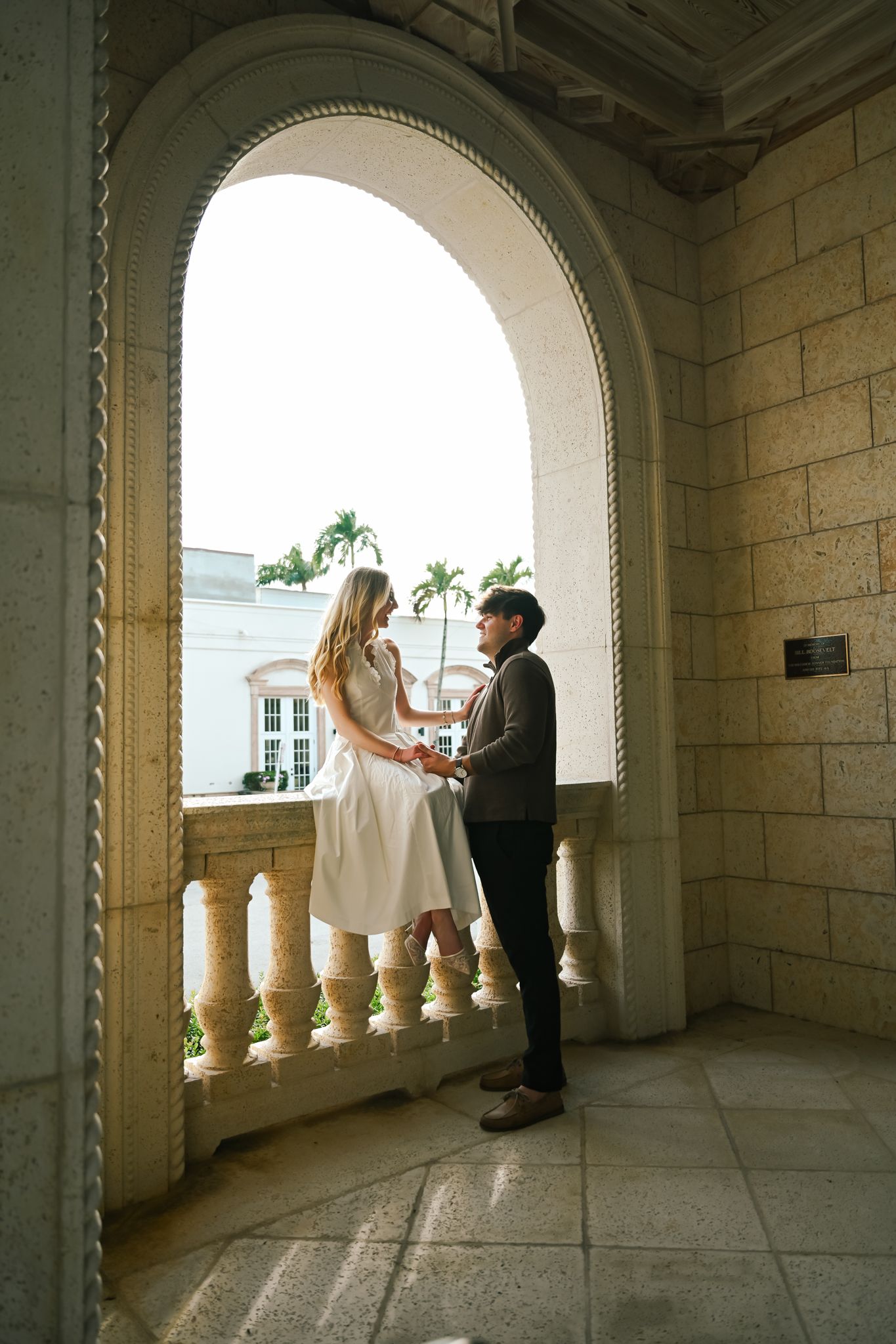 Couple holding hands under a grand stone arch on a balcony with palm trees in the background.