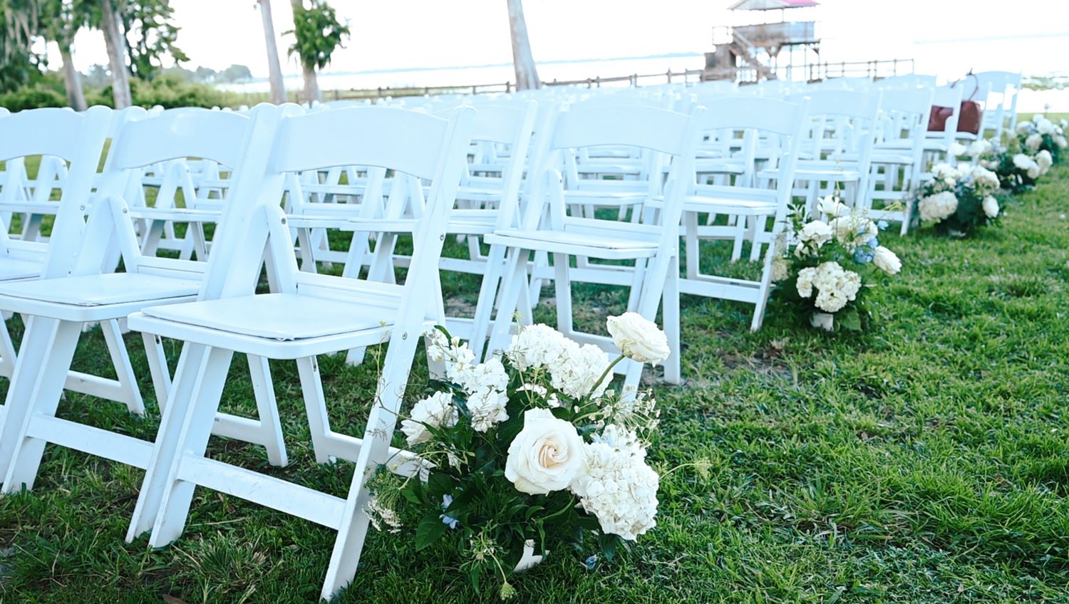 White chairs arranged on grass for an outdoor wedding ceremony with floral decorations.