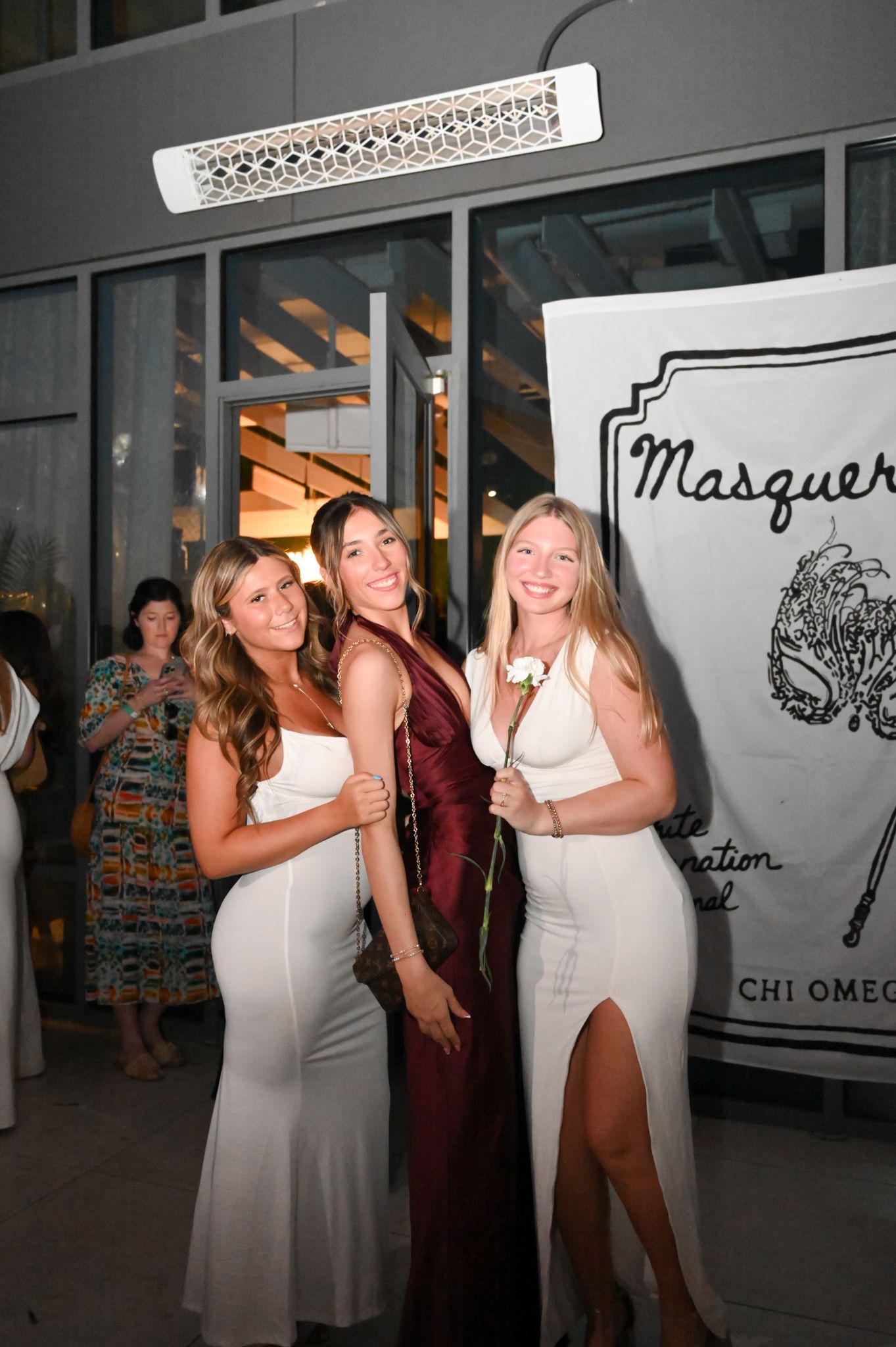 Three young women in formal dresses pose together at a Chi Omega Masquerade event, two in white gowns and one in burgundy.