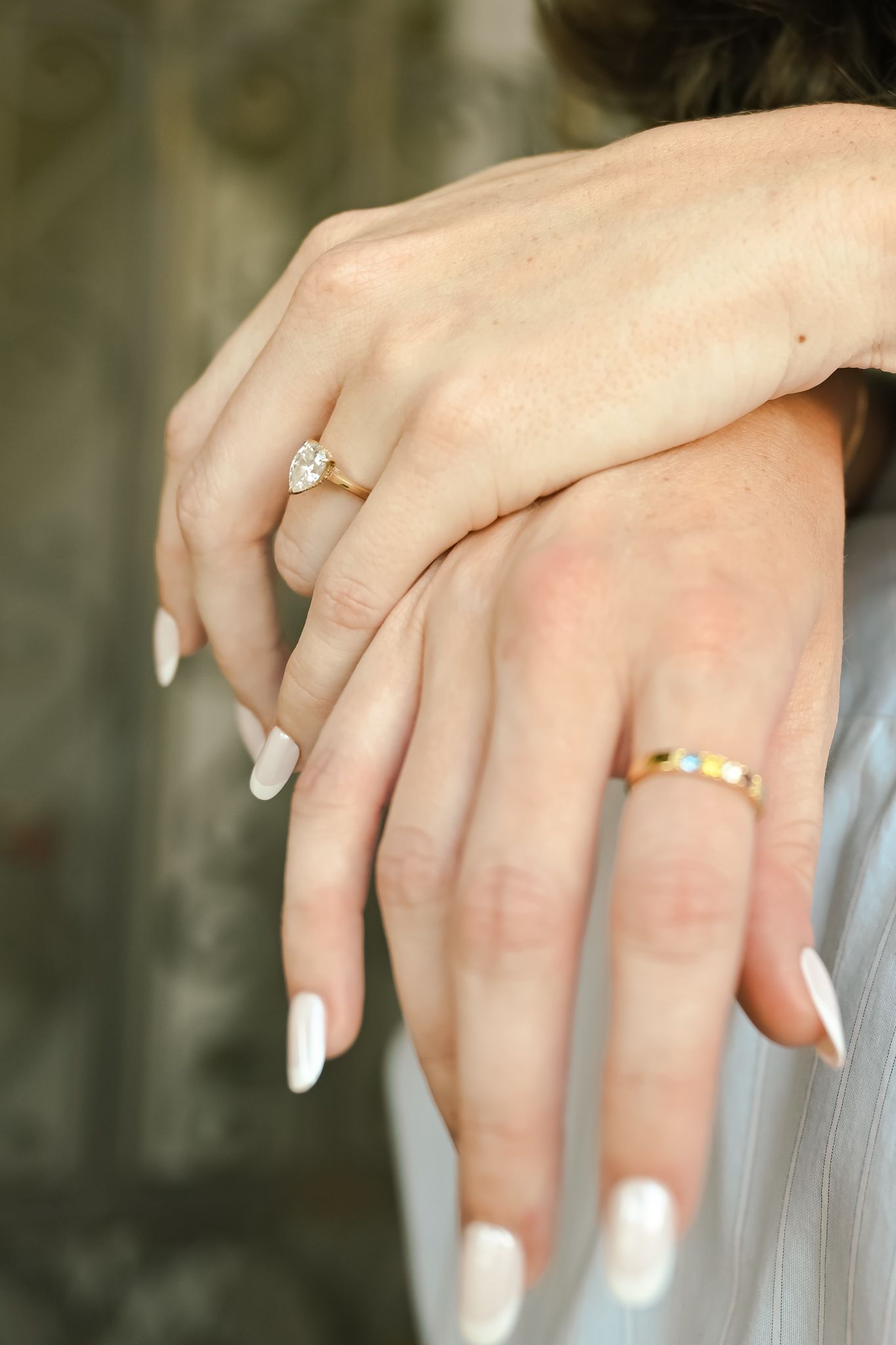 Two hands displaying gold rings, one with a diamond solitaire and one with colorful gems, with white manicured nails.