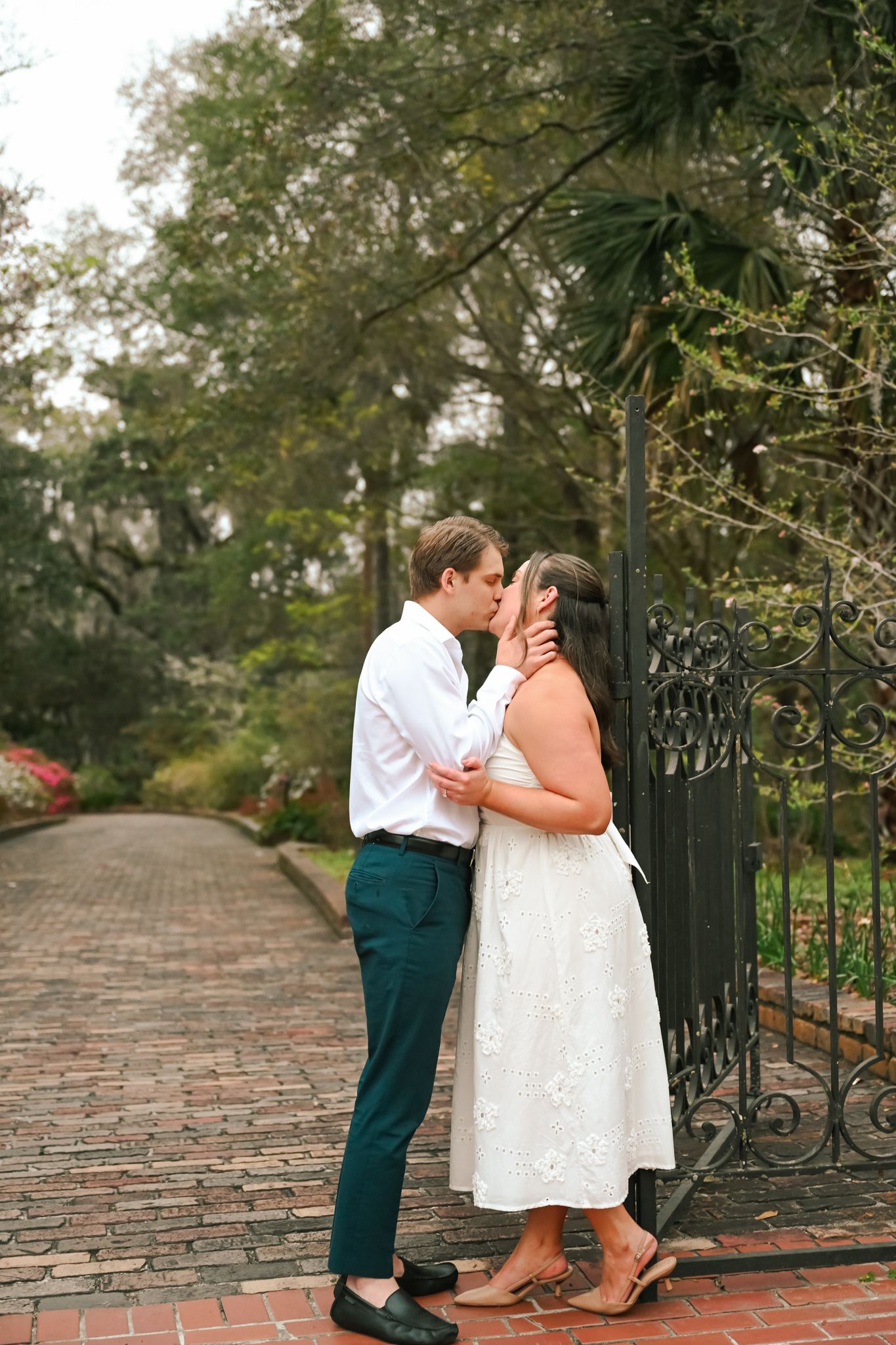 Couple sharing a romantic kiss by an ornate iron gate on a brick path surrounded by lush greenery.