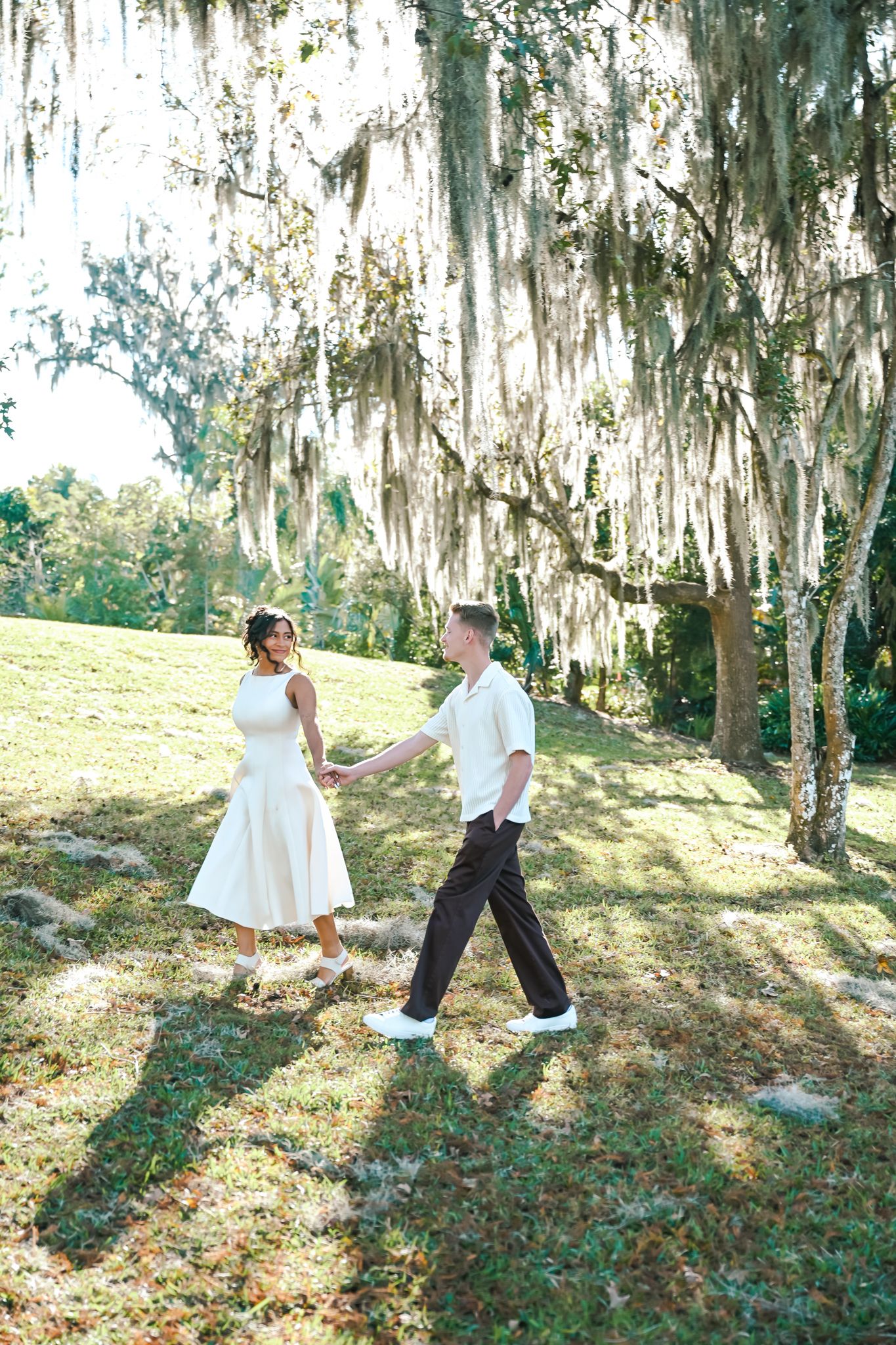 Couple holding hands and dancing under Spanish moss-draped oak trees on a sunny green lawn.