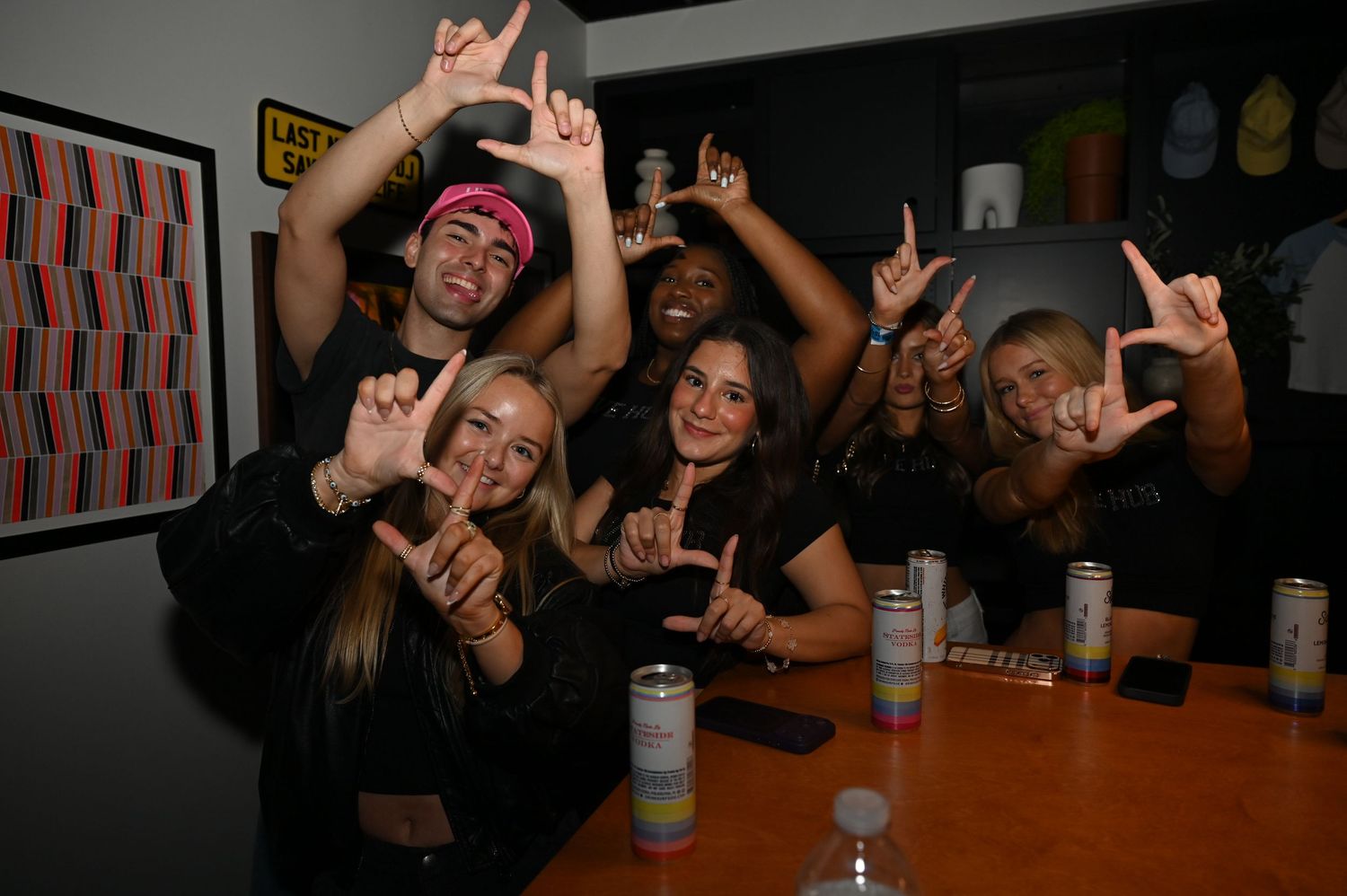 Group of friends posing with hand signs at a party, smiling at camera with drinks on table.