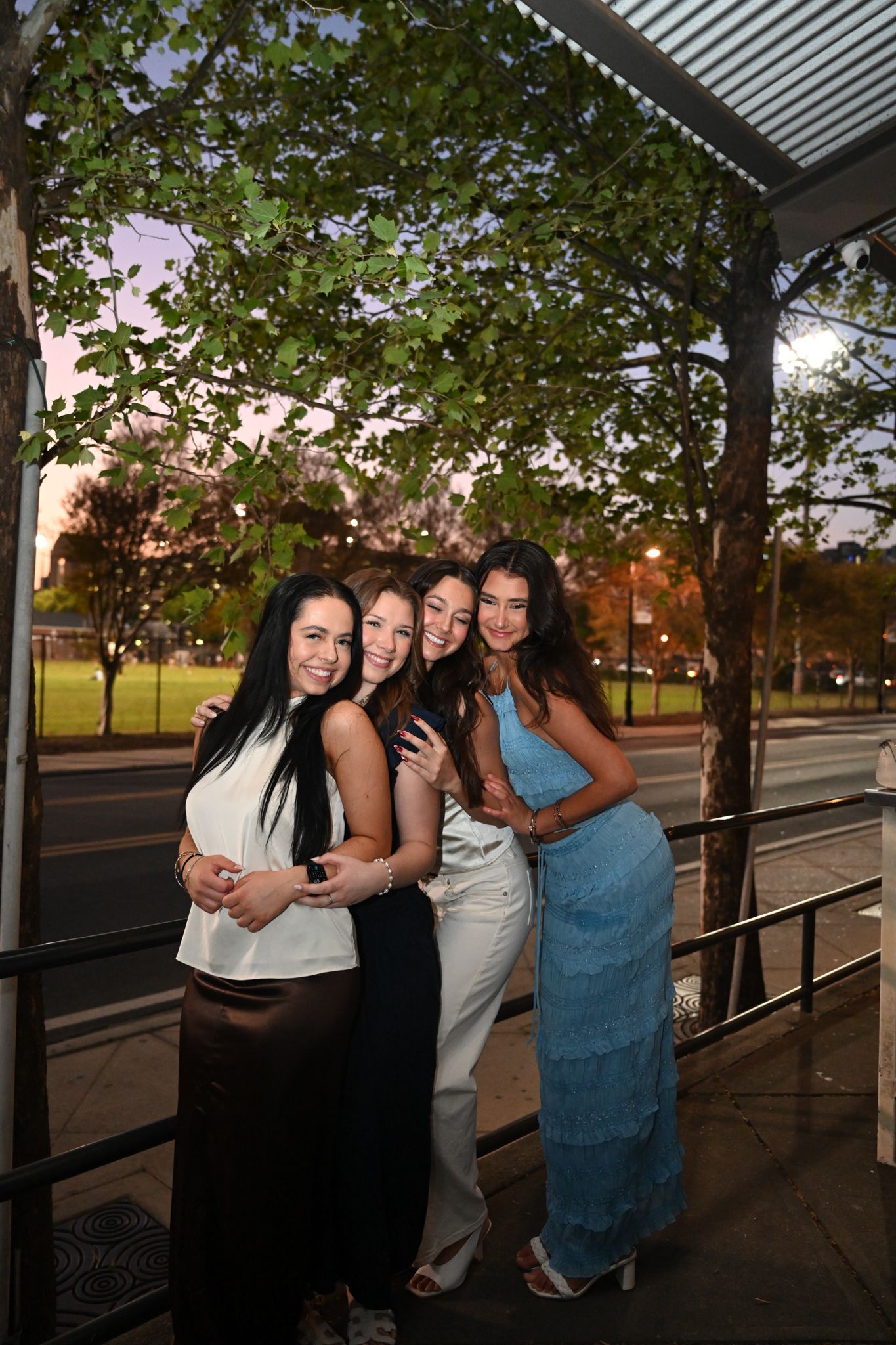 Four young women smiling and posing together under a pergola at dusk outdoors.