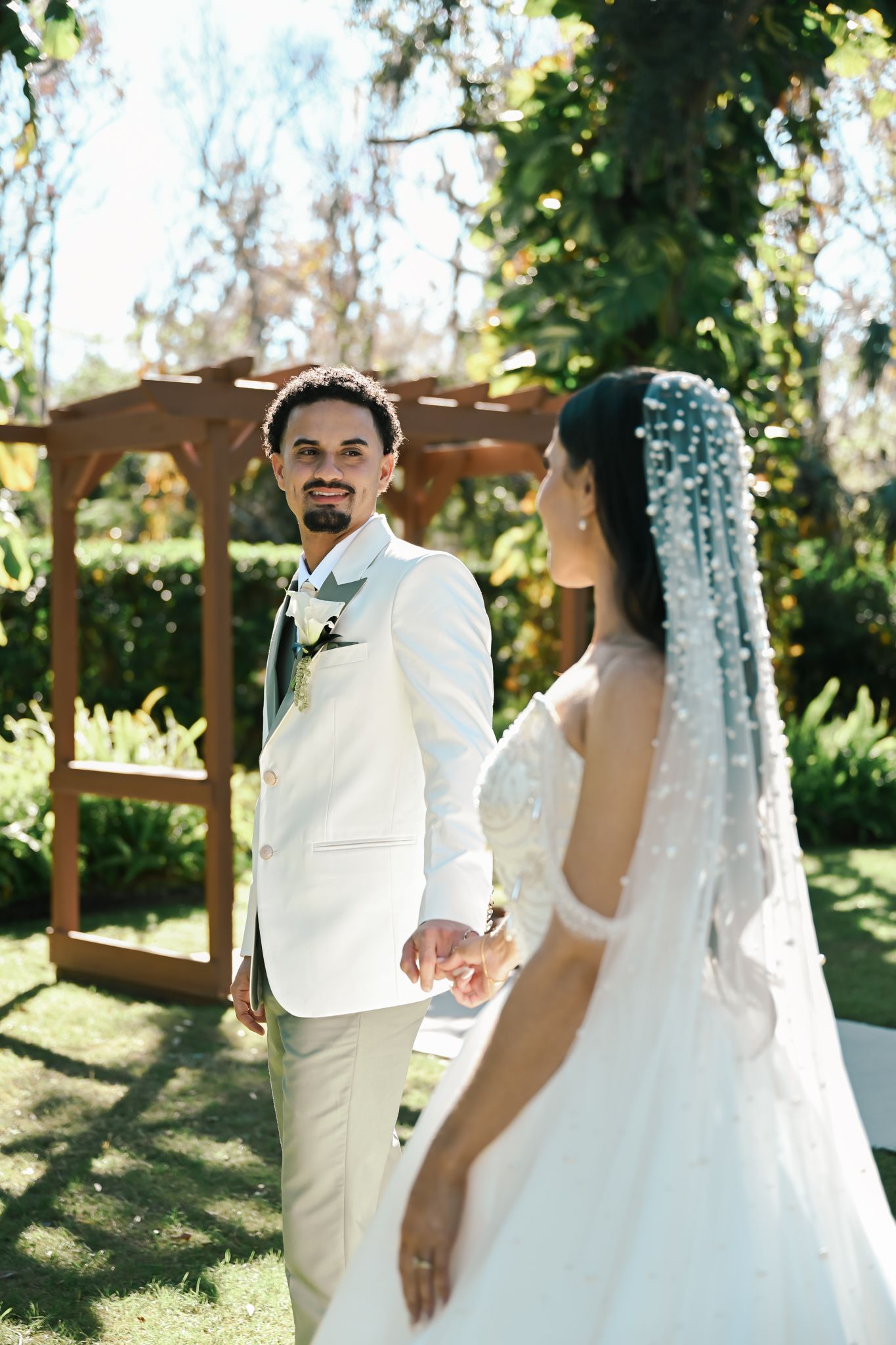 Bride and groom hold hands outdoors at a garden wedding ceremony, groom in white suit, bride in gown with pearl veil.