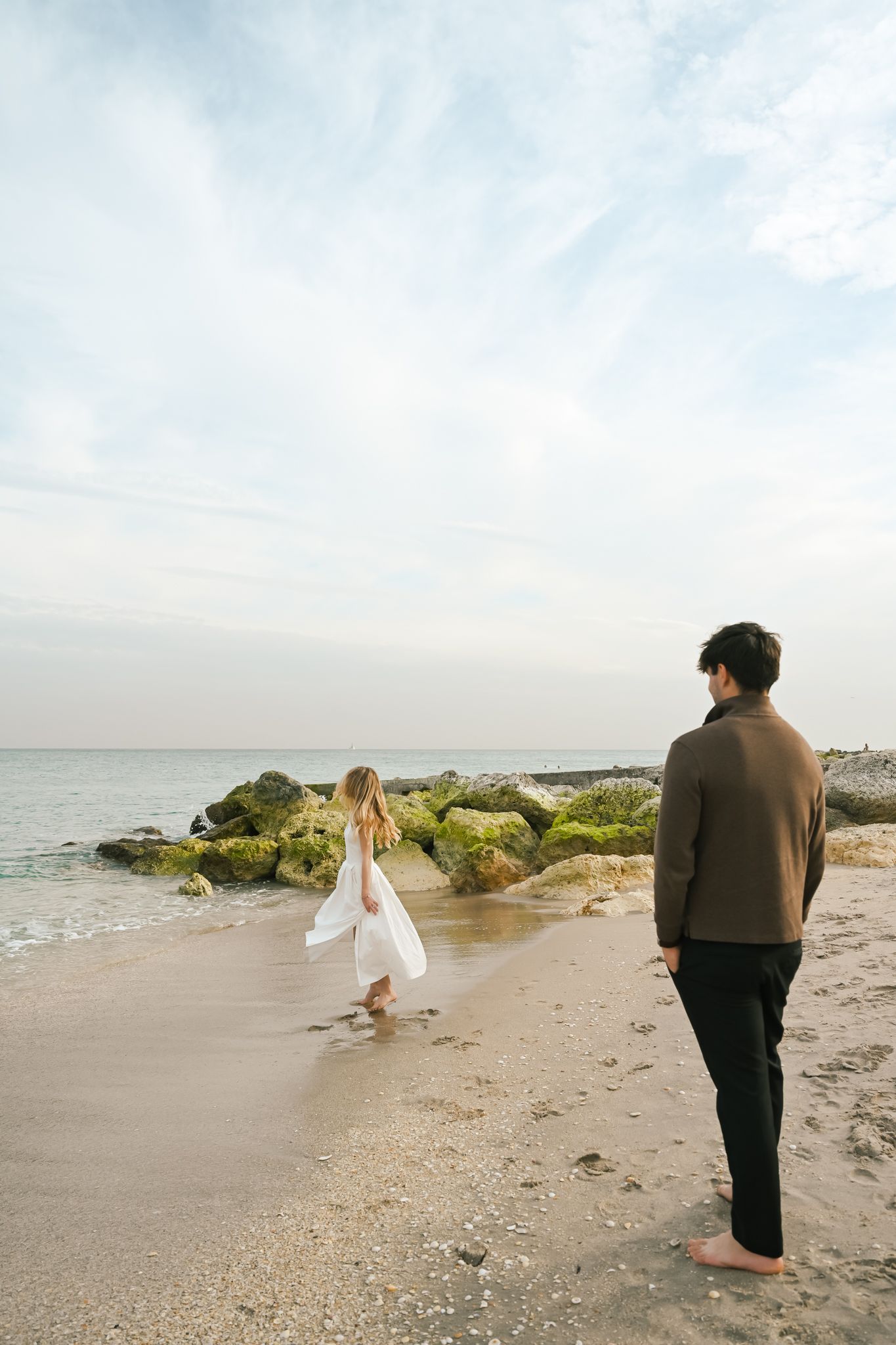 A couple walks barefoot on a rocky beach at dusk, woman in white dress and man in brown jacket.