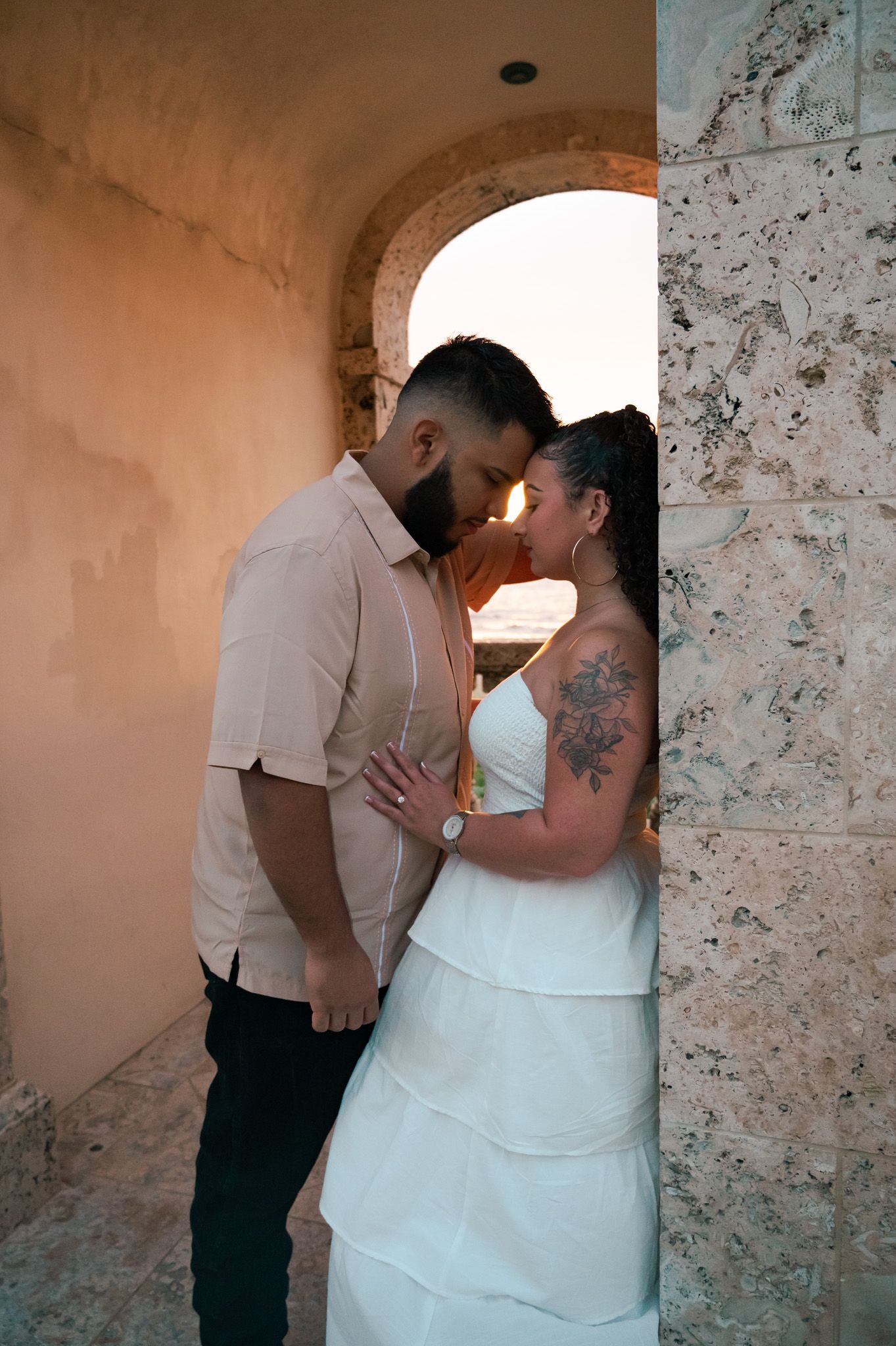 Couple sharing a romantic moment in a stone archway, bride in white dress with floral tattoo, groom in beige shirt.