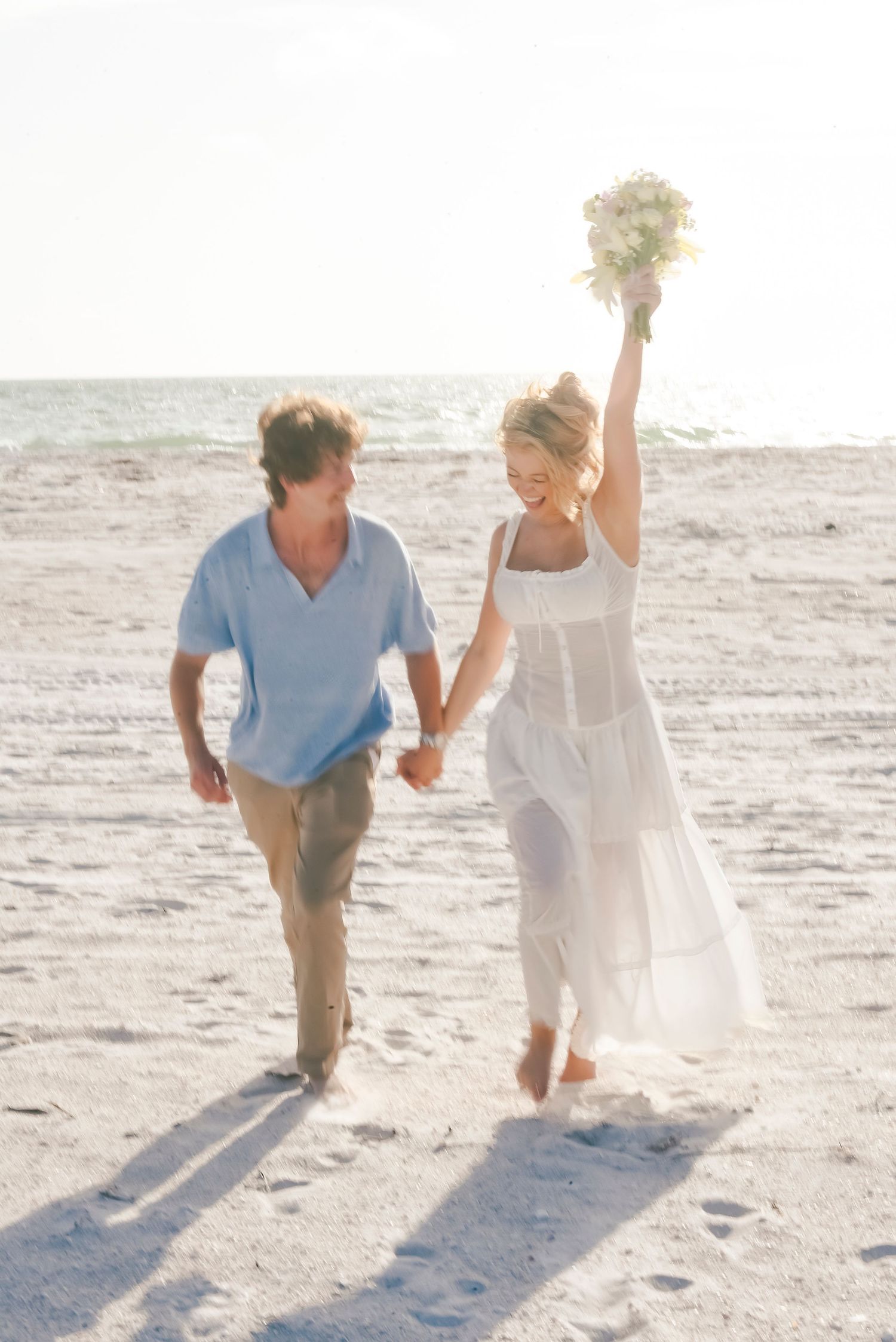 Happy newlyweds walking on a sunny beach, bride in white dress raising bouquet, groom in blue shirt holding hands.