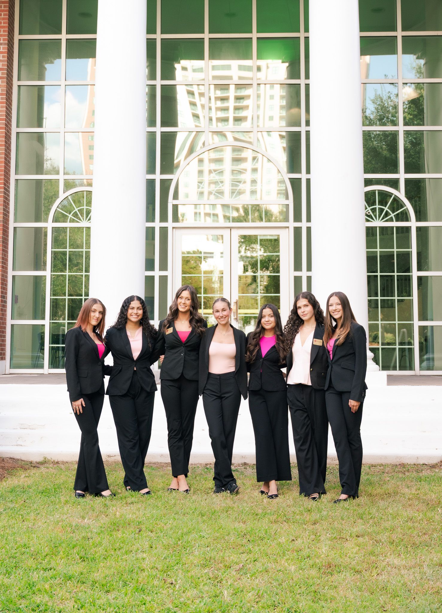 Seven women in matching black suits with pink tops pose together outdoors in front of a white columned building.