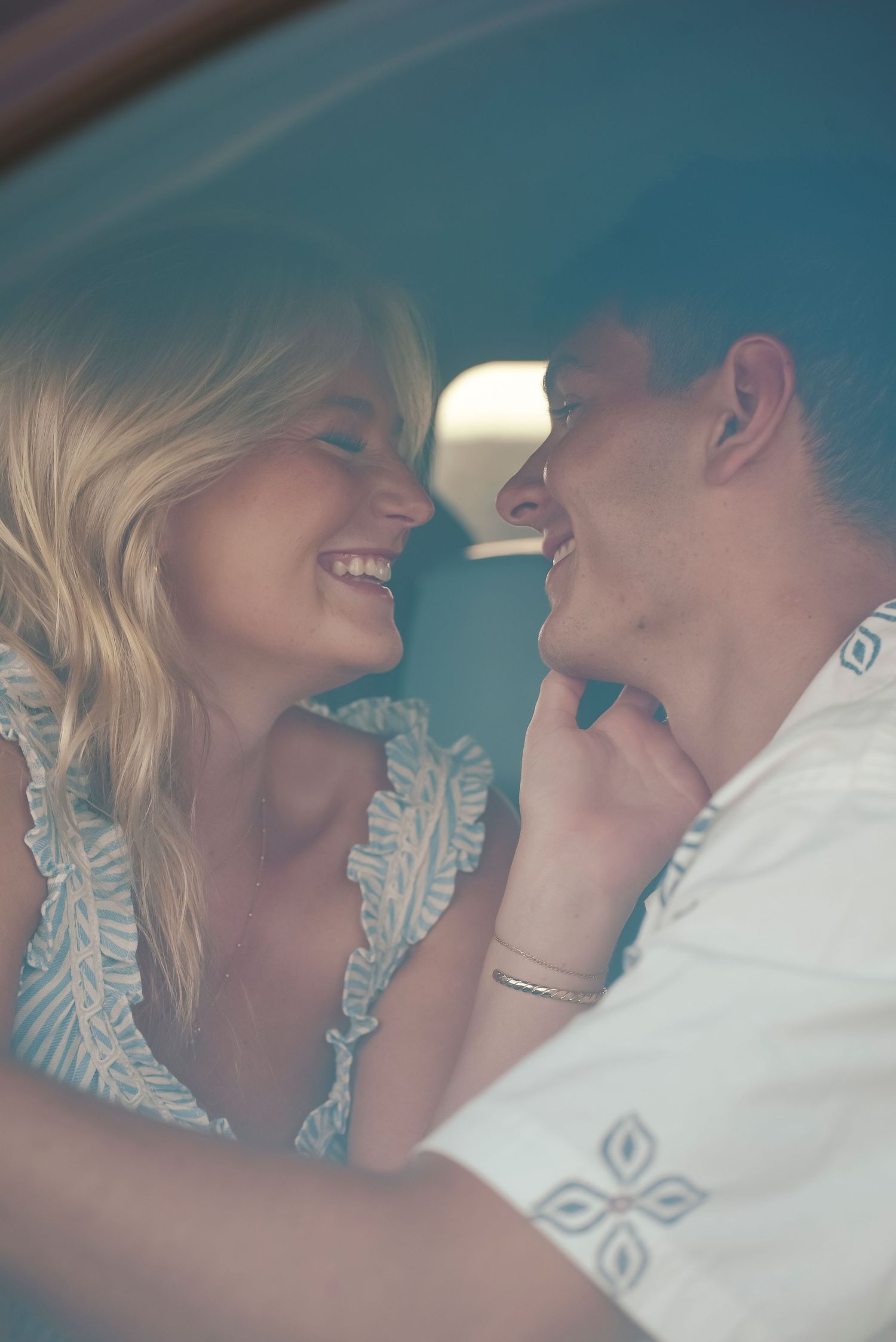 A smiling blonde woman and a man share a romantic moment together inside a car, gazing at each other closely.
