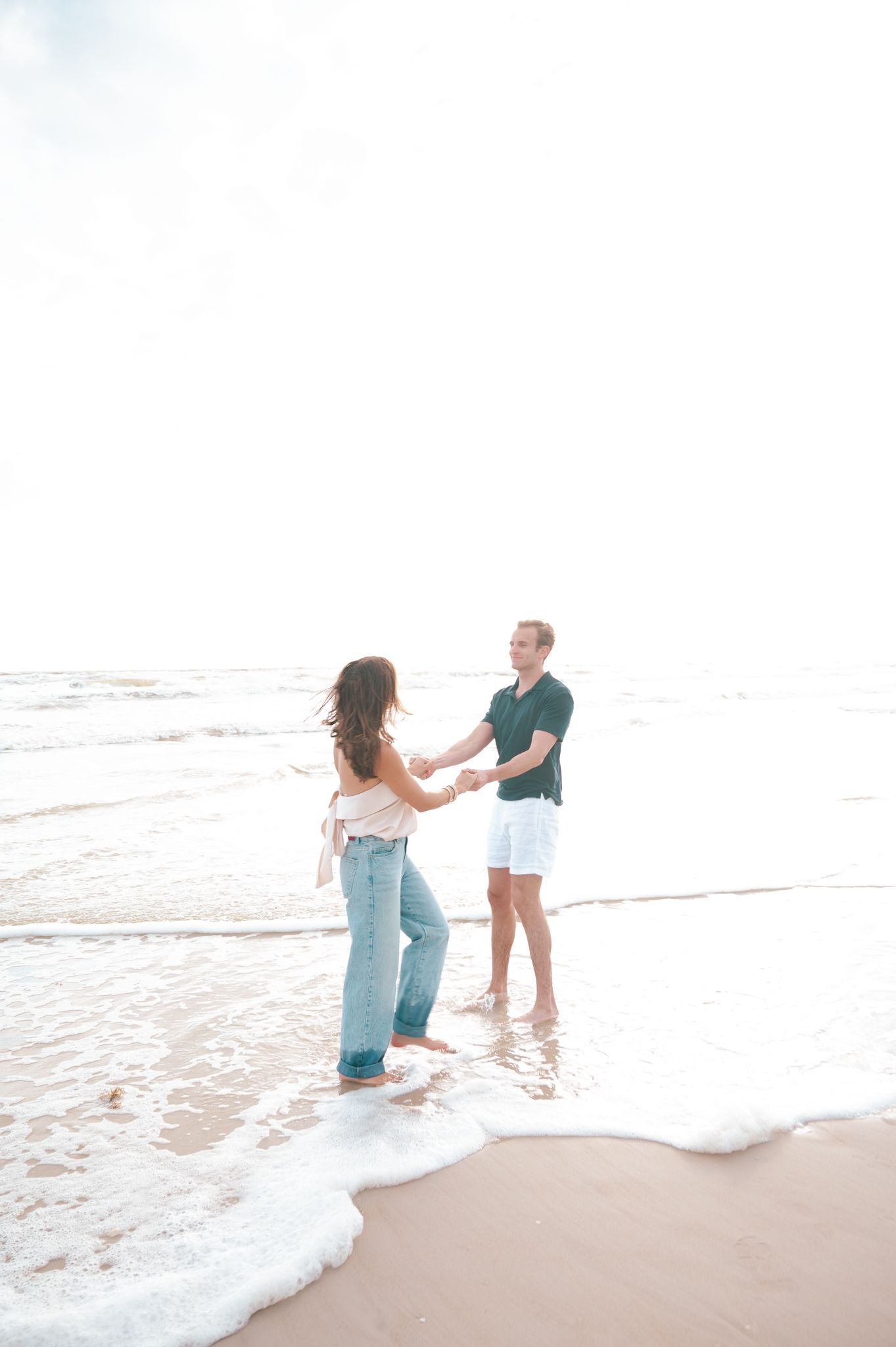 Couple holding hands and dancing together at the beach shoreline, waves washing over their feet.