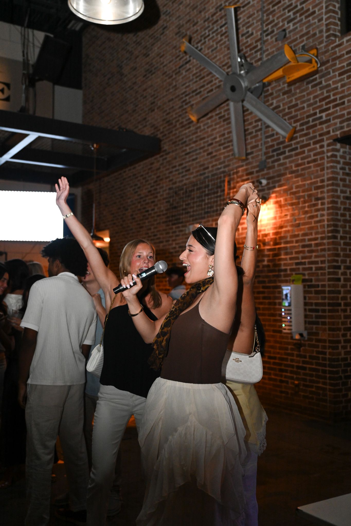 Two women singing karaoke with microphone, arms raised, in a brick-walled venue during a lively party event.