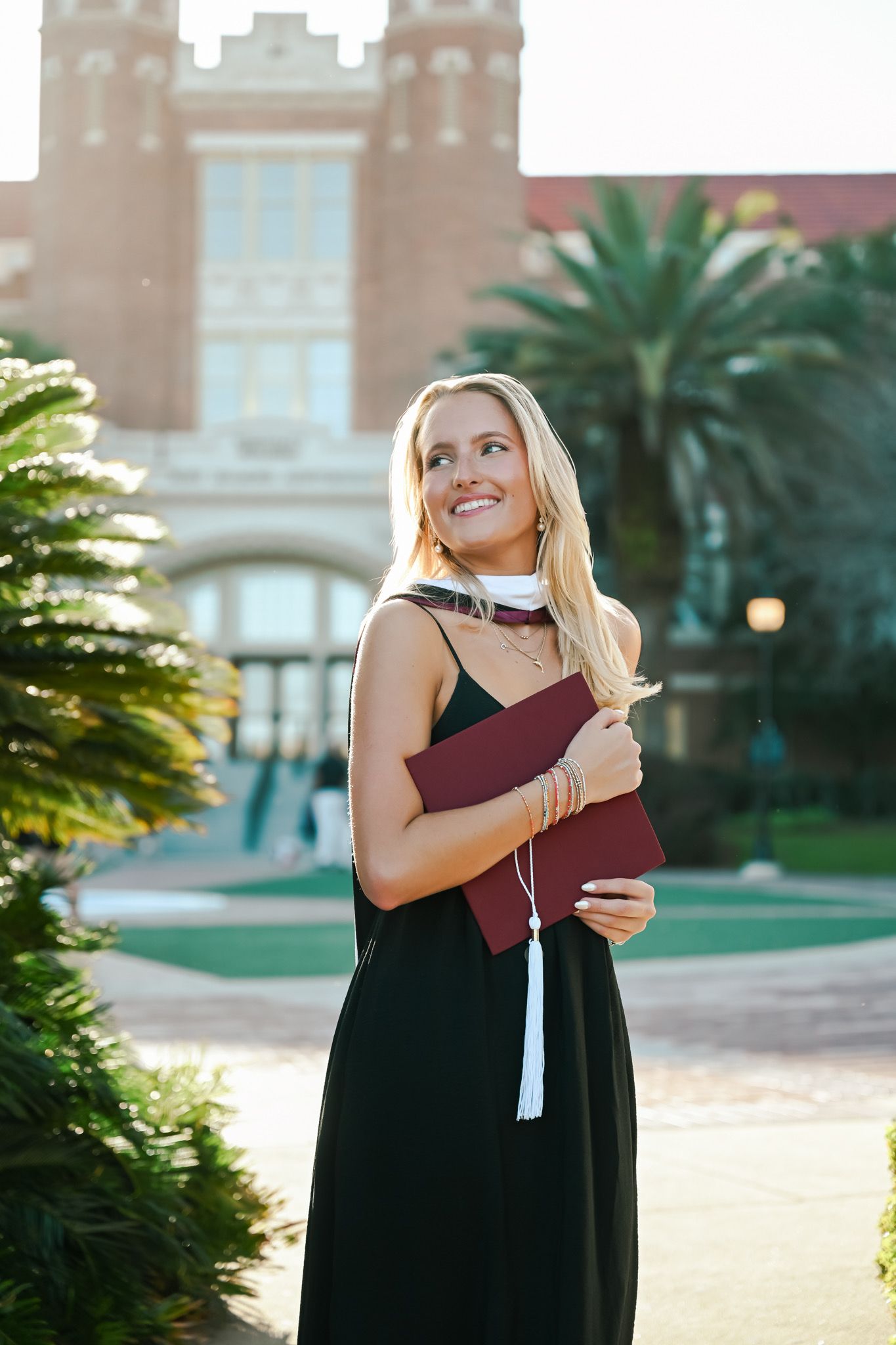 Smiling blonde woman in black dress holding maroon graduation cap outside a university building with palm trees.