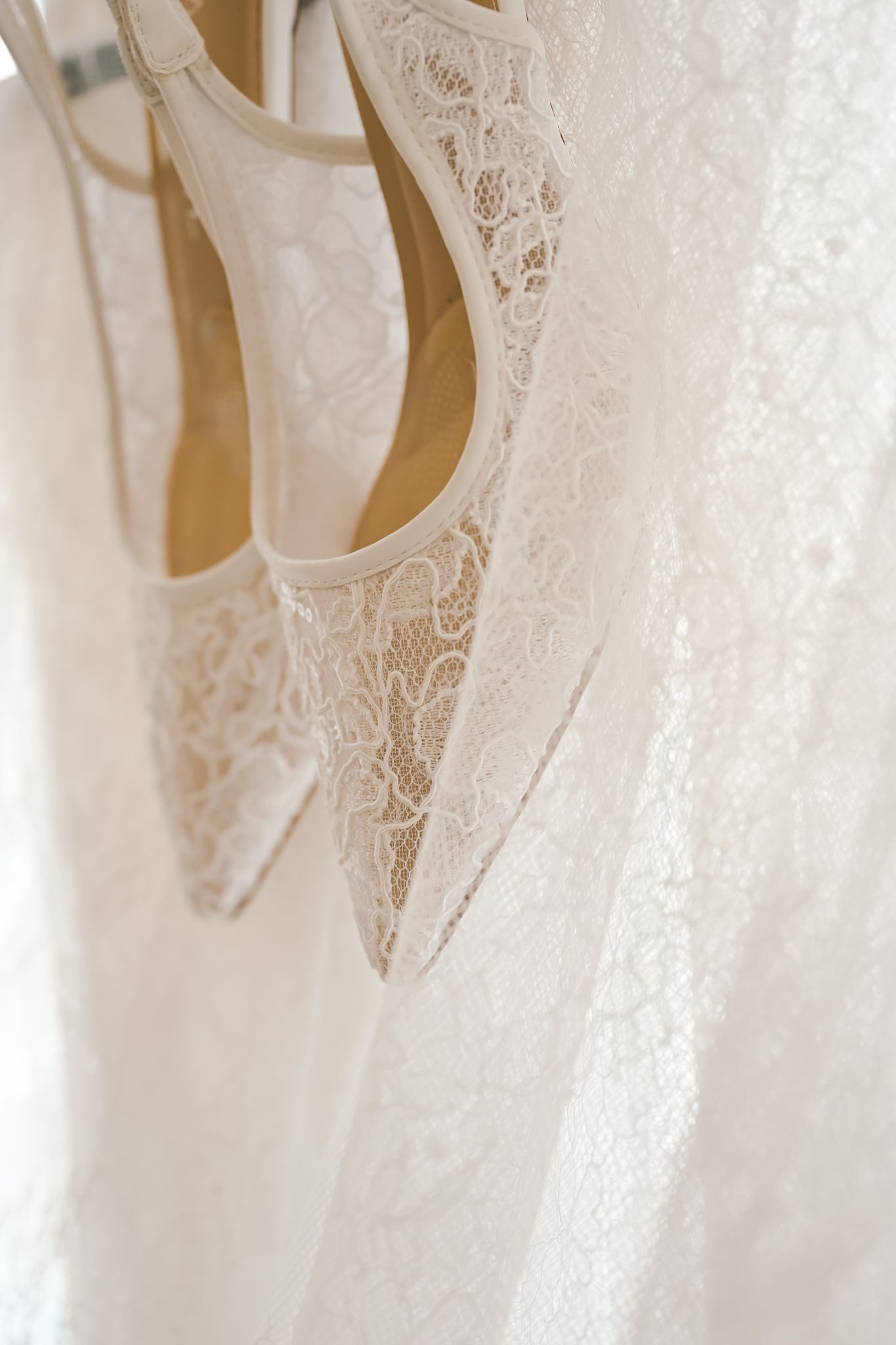 White lace bridal heels hanging against a flowing lace wedding dress, elegant wedding day accessories.