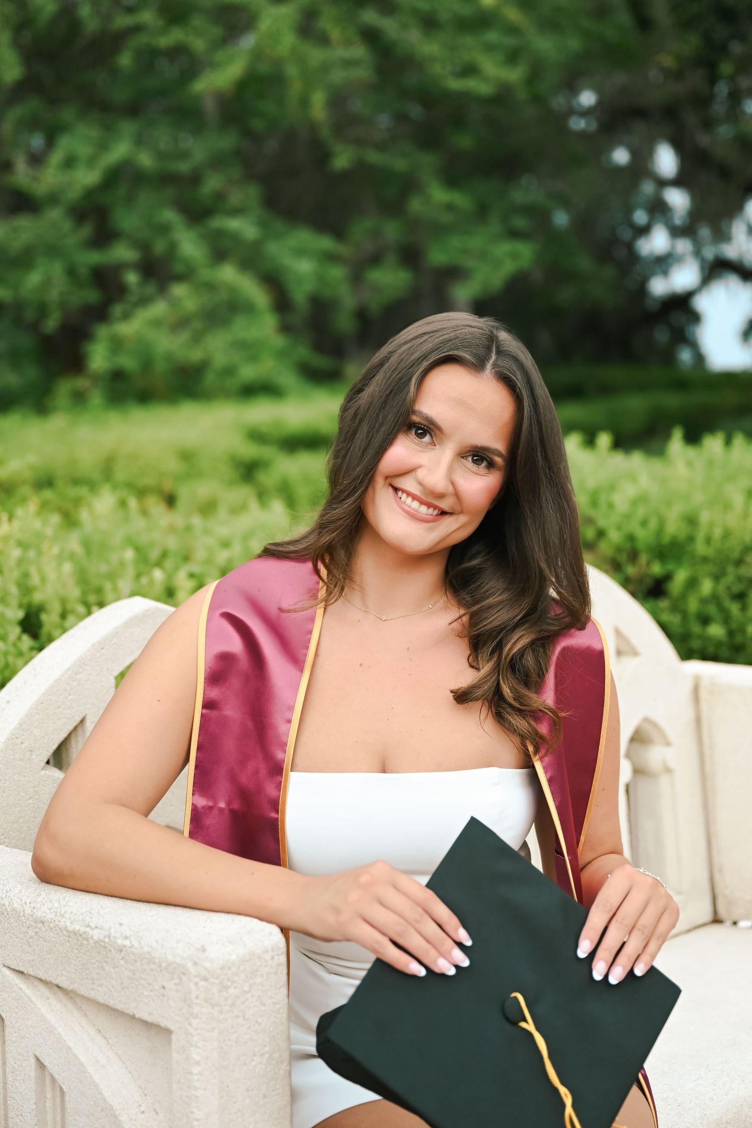 Smiling young woman in white dress with maroon graduation stole holds cap on white bench outdoors.