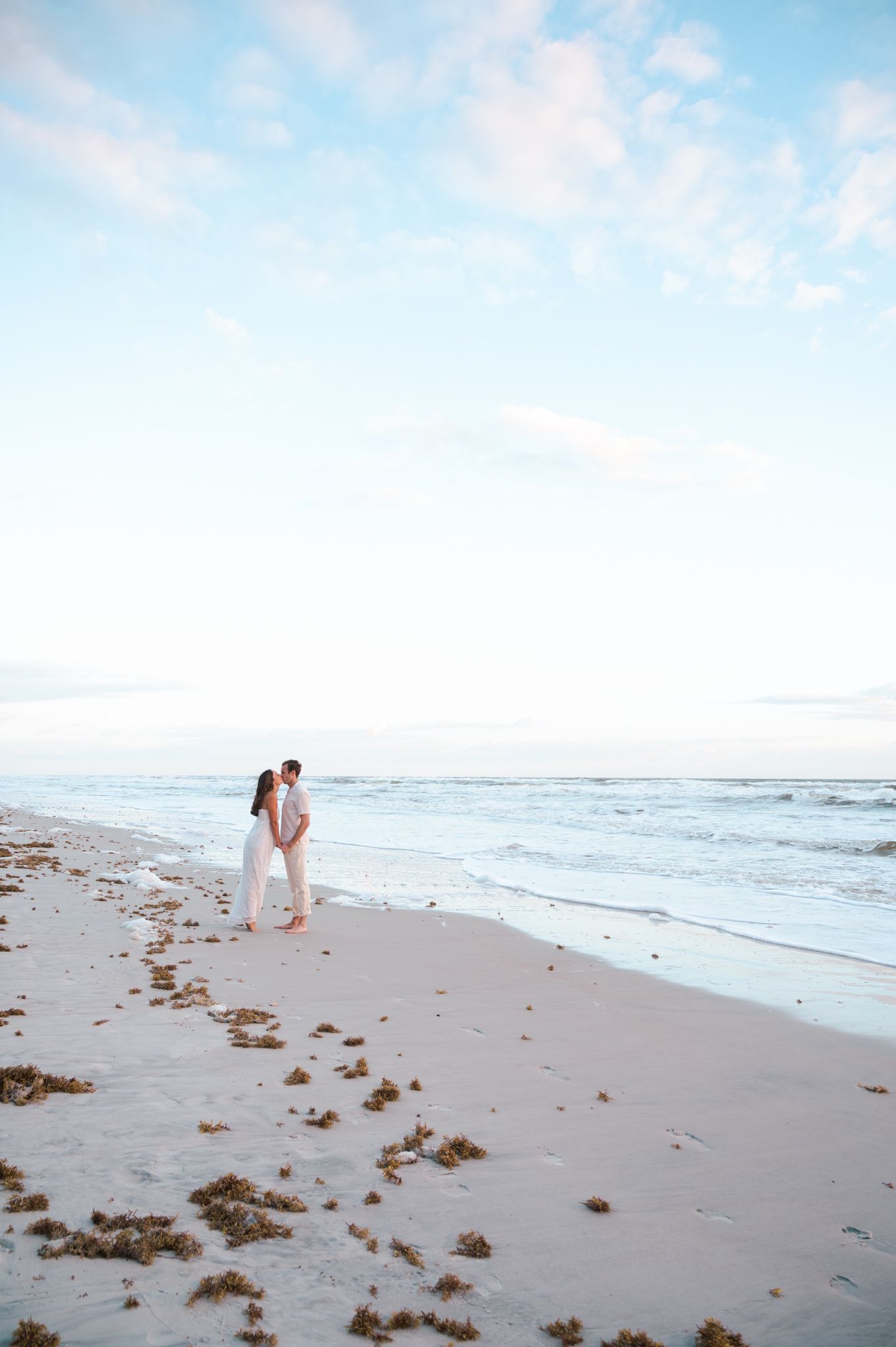 Couple embracing on a serene white sand beach at dusk with gentle ocean waves and a soft pastel sky.