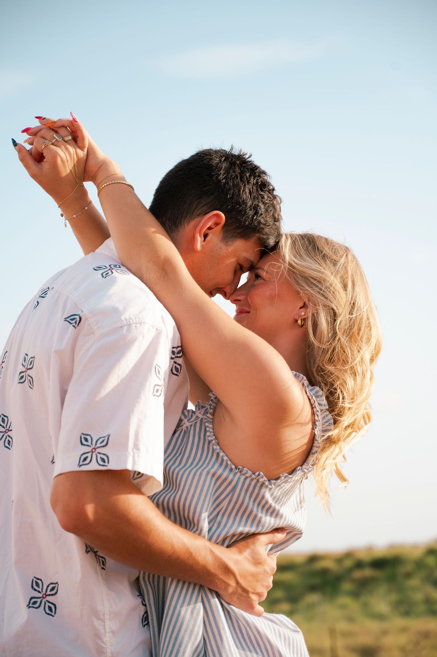 A romantic couple sharing an intimate moment outdoors, with the woman raising their hands while embracing under a clear blue sky.