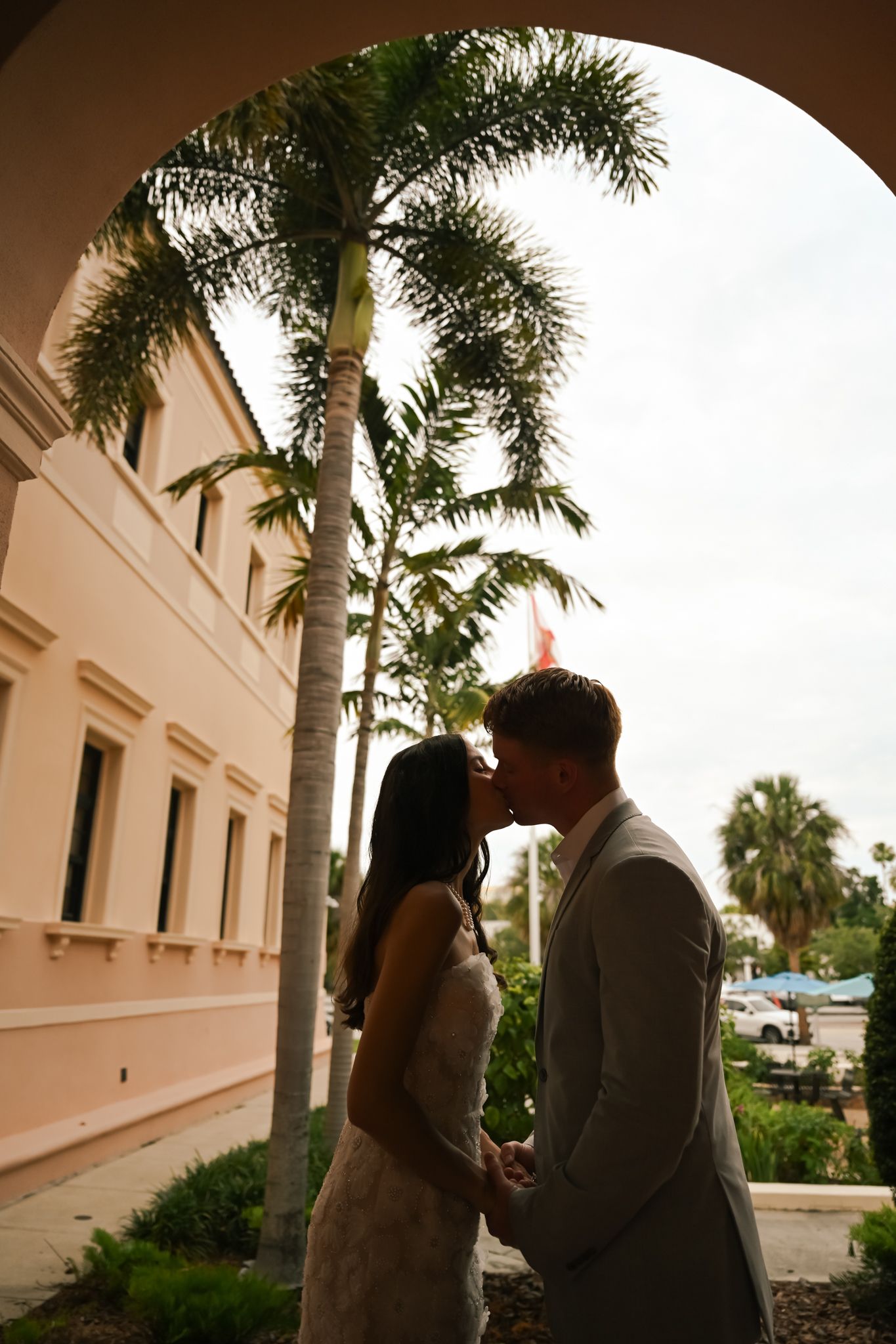 Couple sharing a romantic kiss in silhouette under an arch, with tall palm trees and a pink building backdrop.