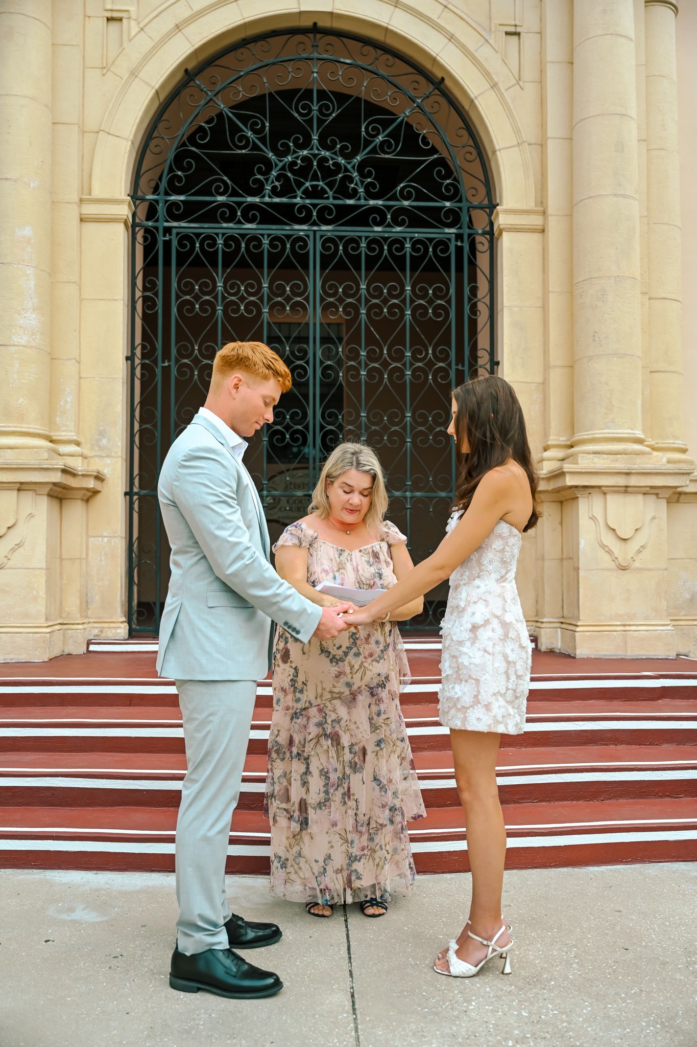 Couple holding hands during outdoor wedding ceremony before ornate church doors with officiant.