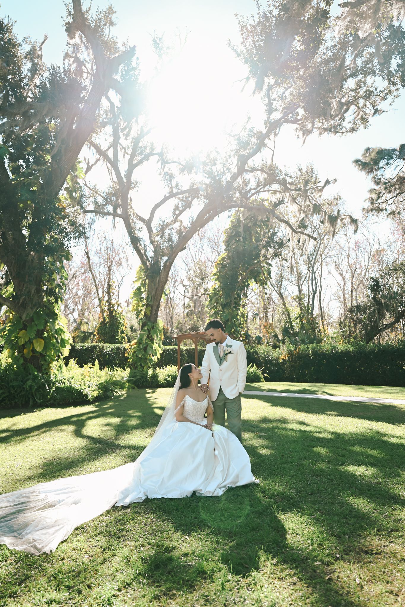 Couple in wedding attire sitting together on grassy hillside with sunlight streaming through trees in romantic outdoor setting.