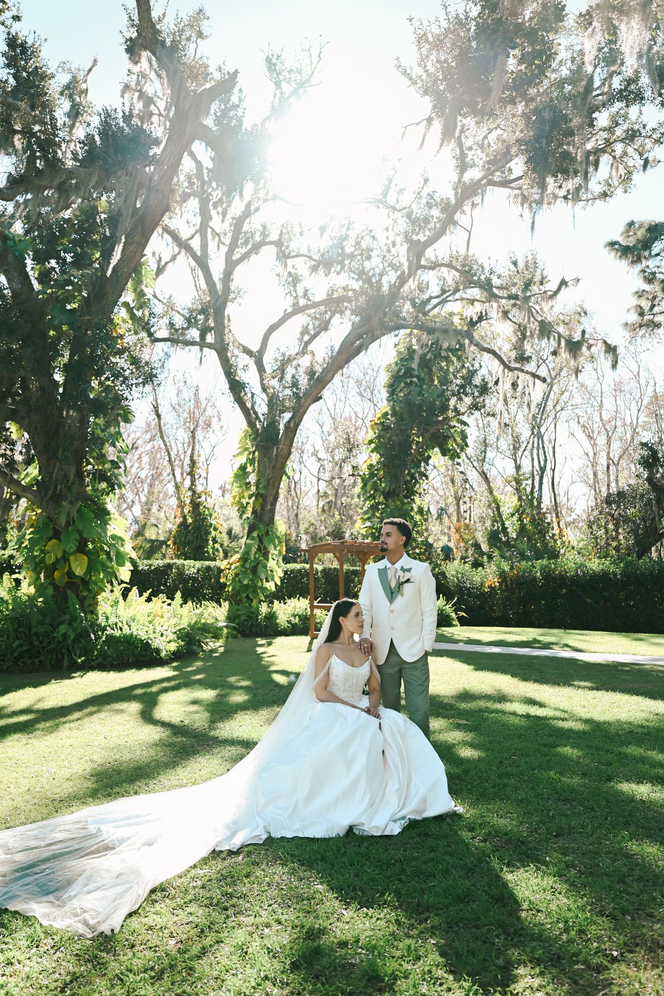 Bride in white gown sits on lush green lawn as groom in tan suit stands beside her under sunlit oak trees.