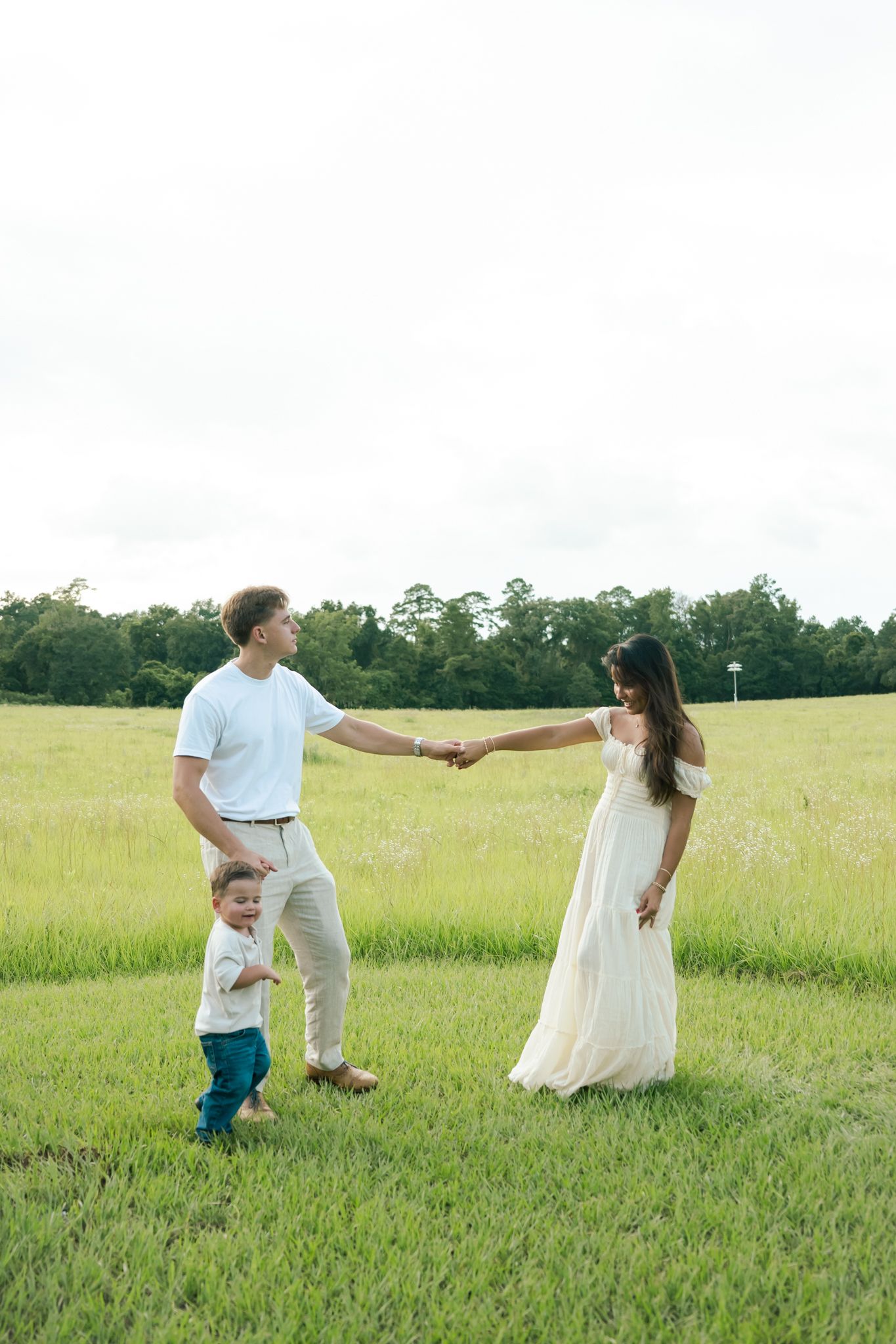 Family of three holding hands in a green field, parents in white attire with toddler, trees in background.