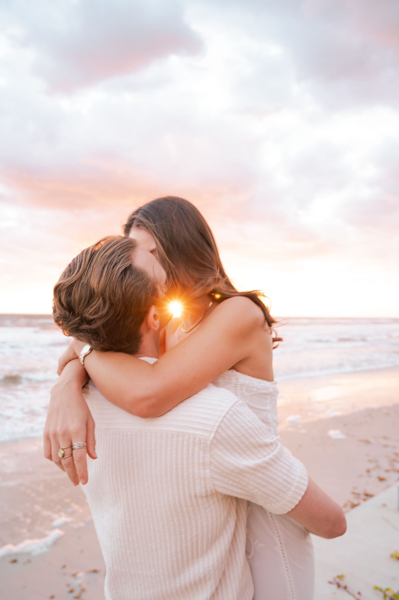 Couple sharing a romantic kiss on the beach at sunset, with golden light and pastel clouds over the ocean.