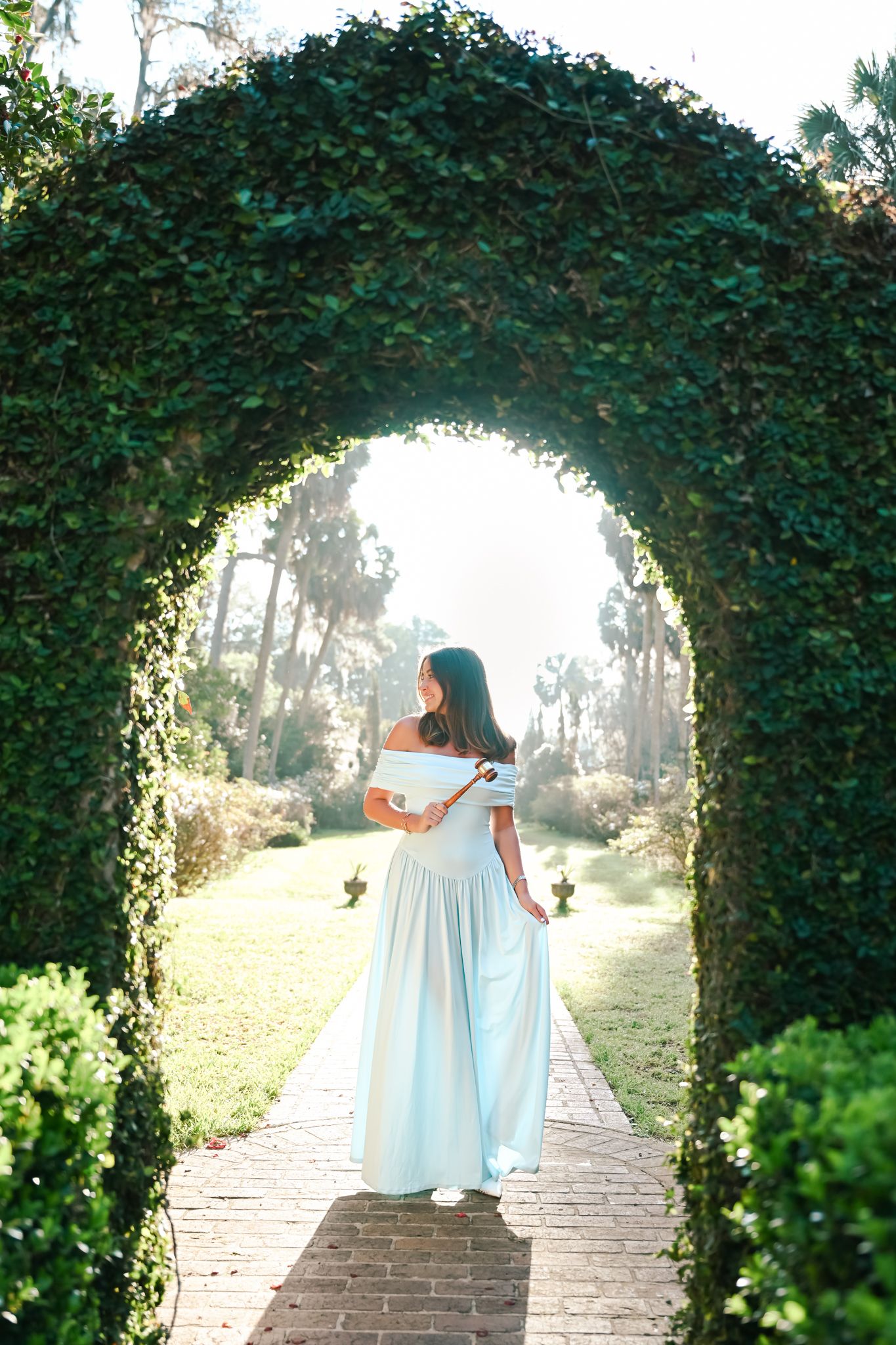 Woman in light blue off-shoulder gown standing under lush green hedge arch on brick pathway in garden.