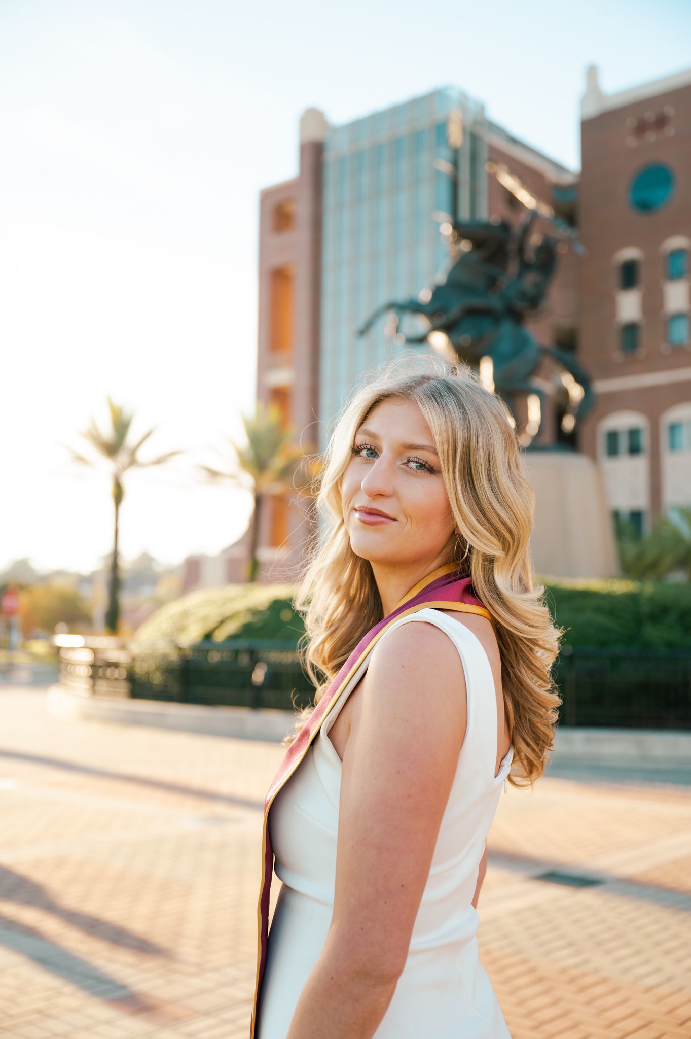 Woman in white top poses against urban backdrop with brick buildings during golden hour sunset.