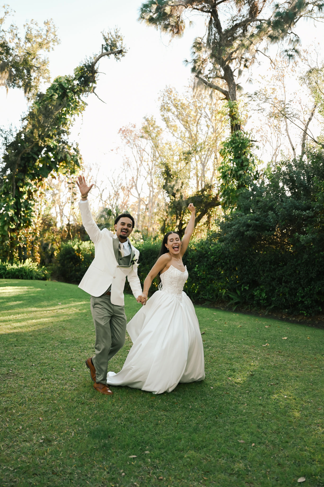 Joyful bride and groom celebrating on green lawn, raising arms in excitement, surrounded by lush trees at outdoor wedding.