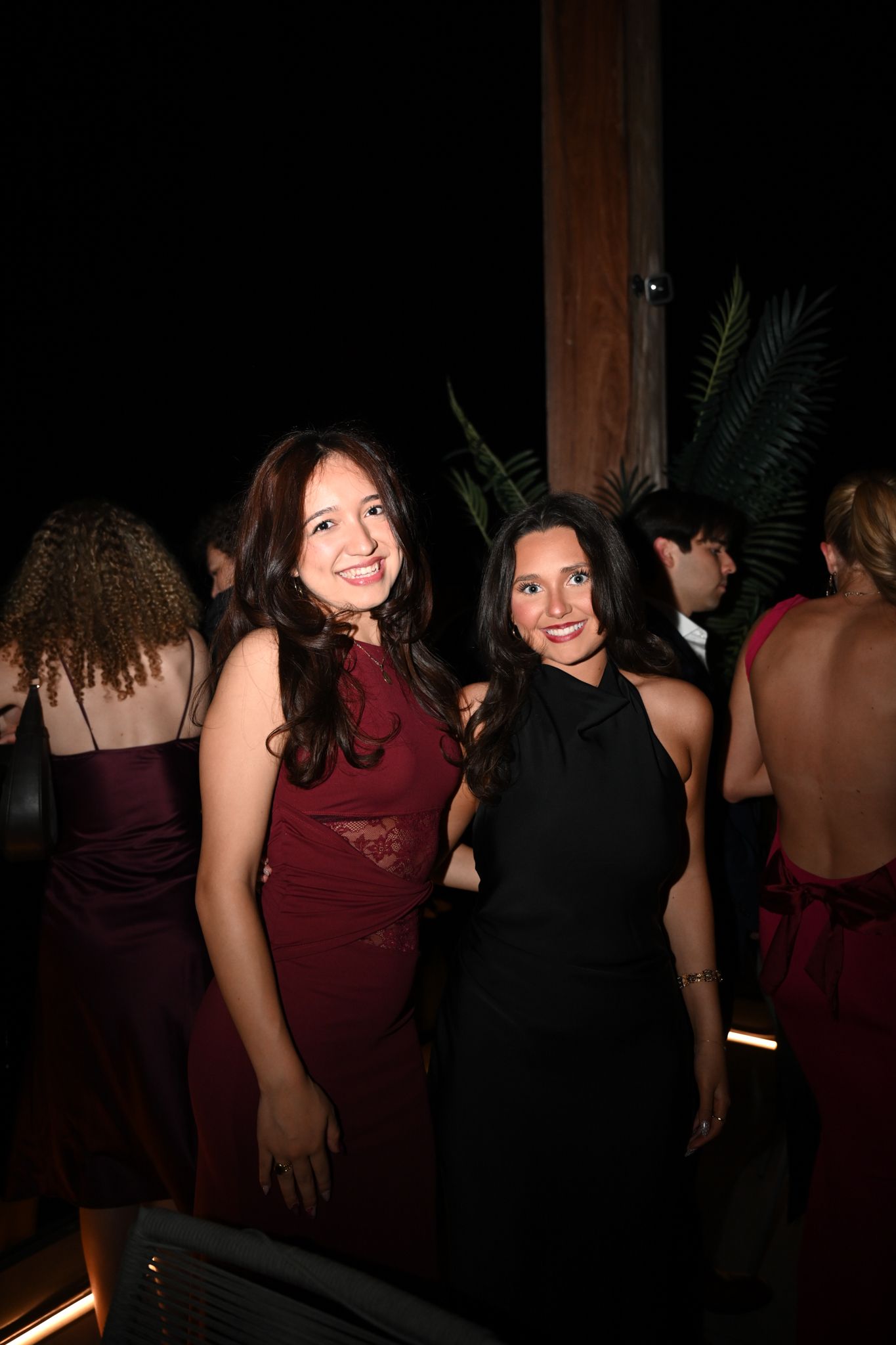Two women in elegant dresses, one burgundy and one black, smiling at a formal evening event.