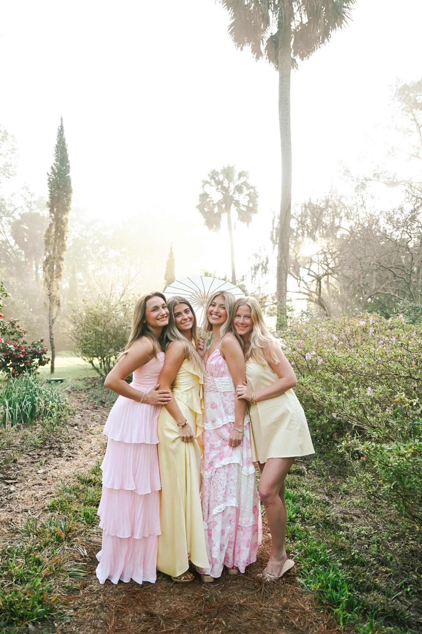 Four women in pastel pink and yellow dresses pose together outdoors among palm trees in soft golden light.