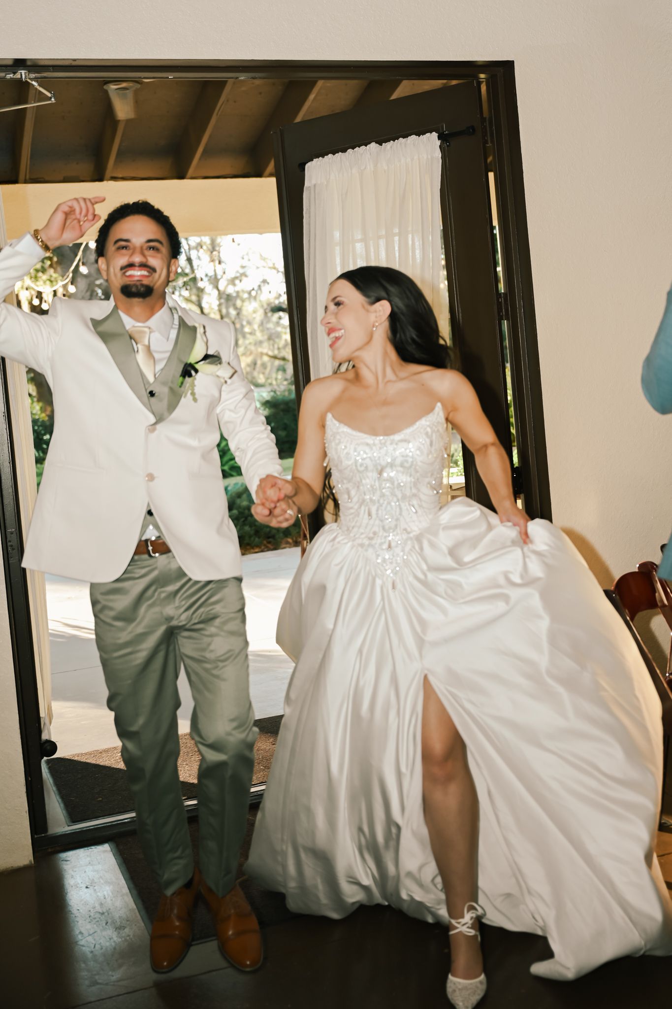 A joyful bride in a white lace gown and groom in sage suit hold hands making a grand entrance at their wedding reception.