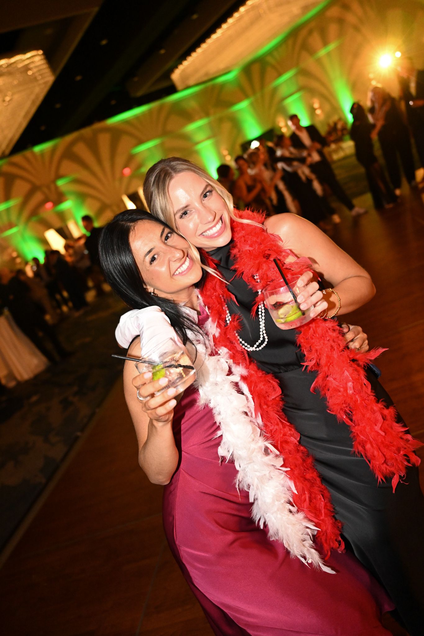 Two women in elegant dresses smile at a formal party, one wearing a red feather boa and pearls, holding cocktails.