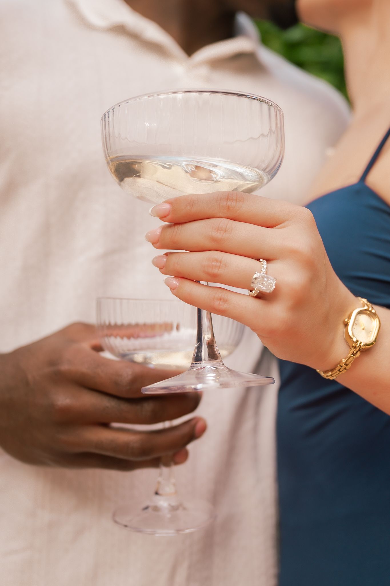 Hands holding champagne glasses with elegant engagement ring and gold jewelry.