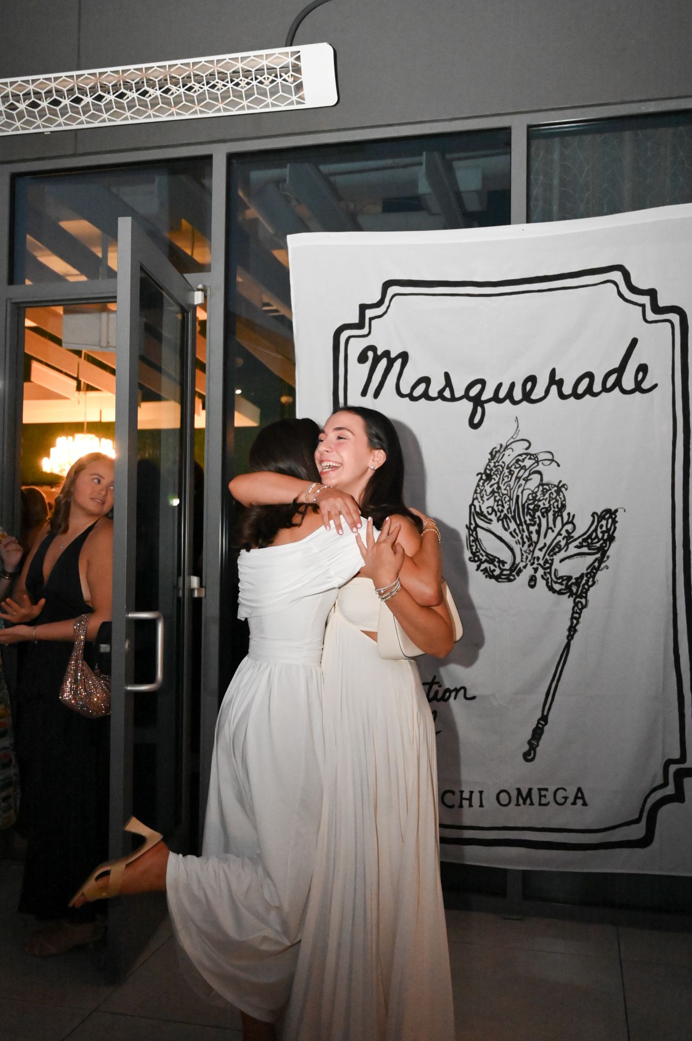 Two women in white dresses embrace at a Chi Omega Masquerade event near an entrance.