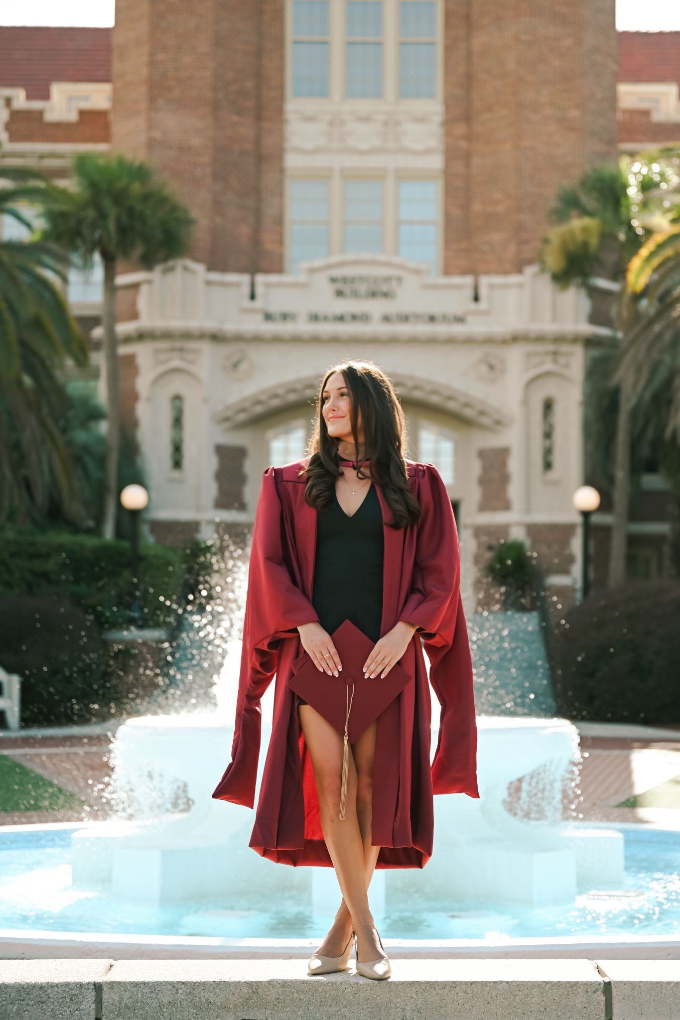 Woman in red graduation gown poses by fountain outside university building.