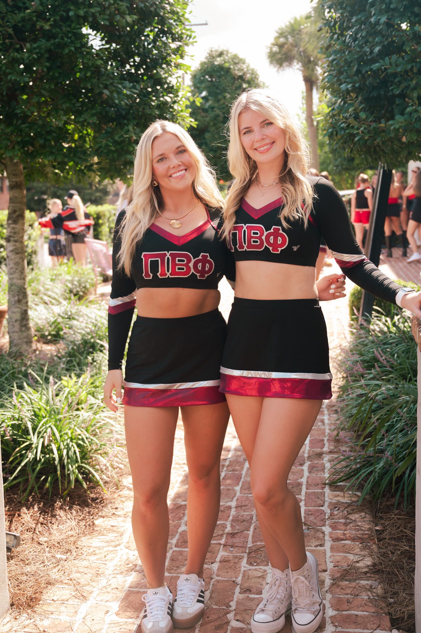 Two blonde women wearing matching Pi Beta Phi sorority cheerleader uniforms smiling outdoors on a brick path.