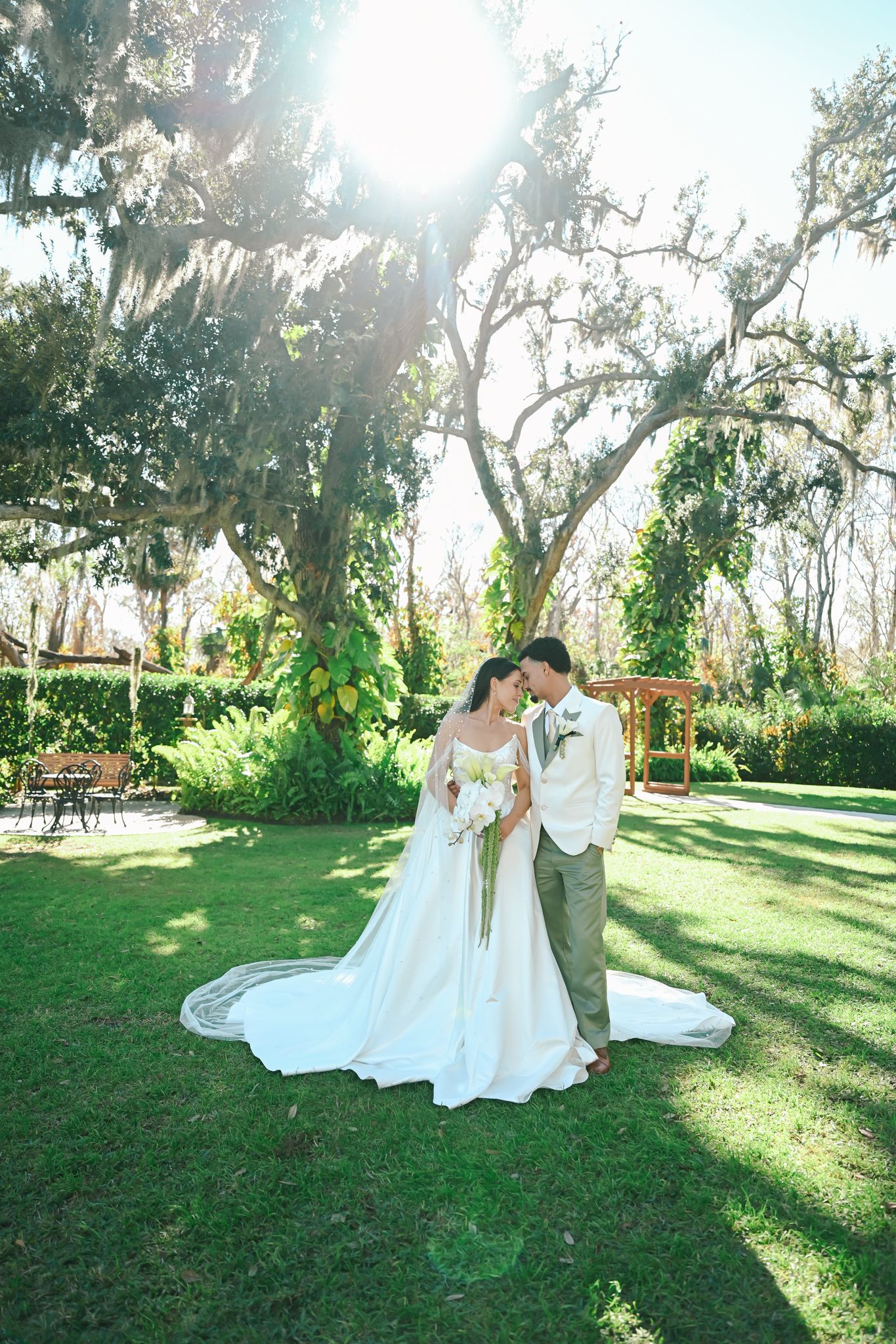 Bride and groom share a romantic moment under a sunlit oak tree in a lush green garden, bride in elegant white gown.