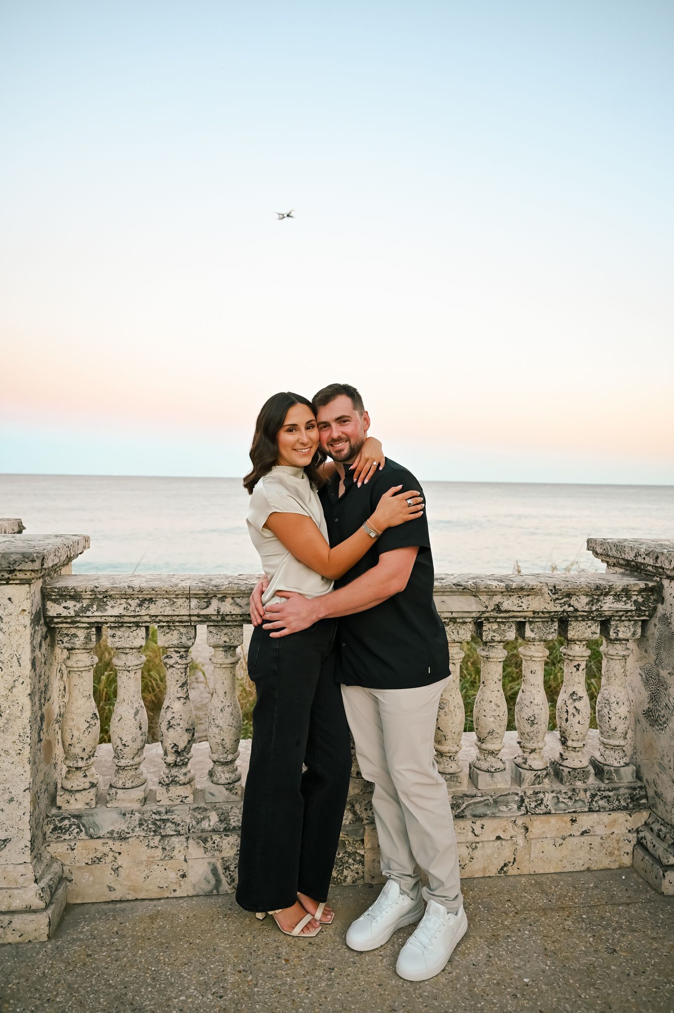 A couple embraces and smiles on a stone balustrade terrace overlooking the ocean at sunset.