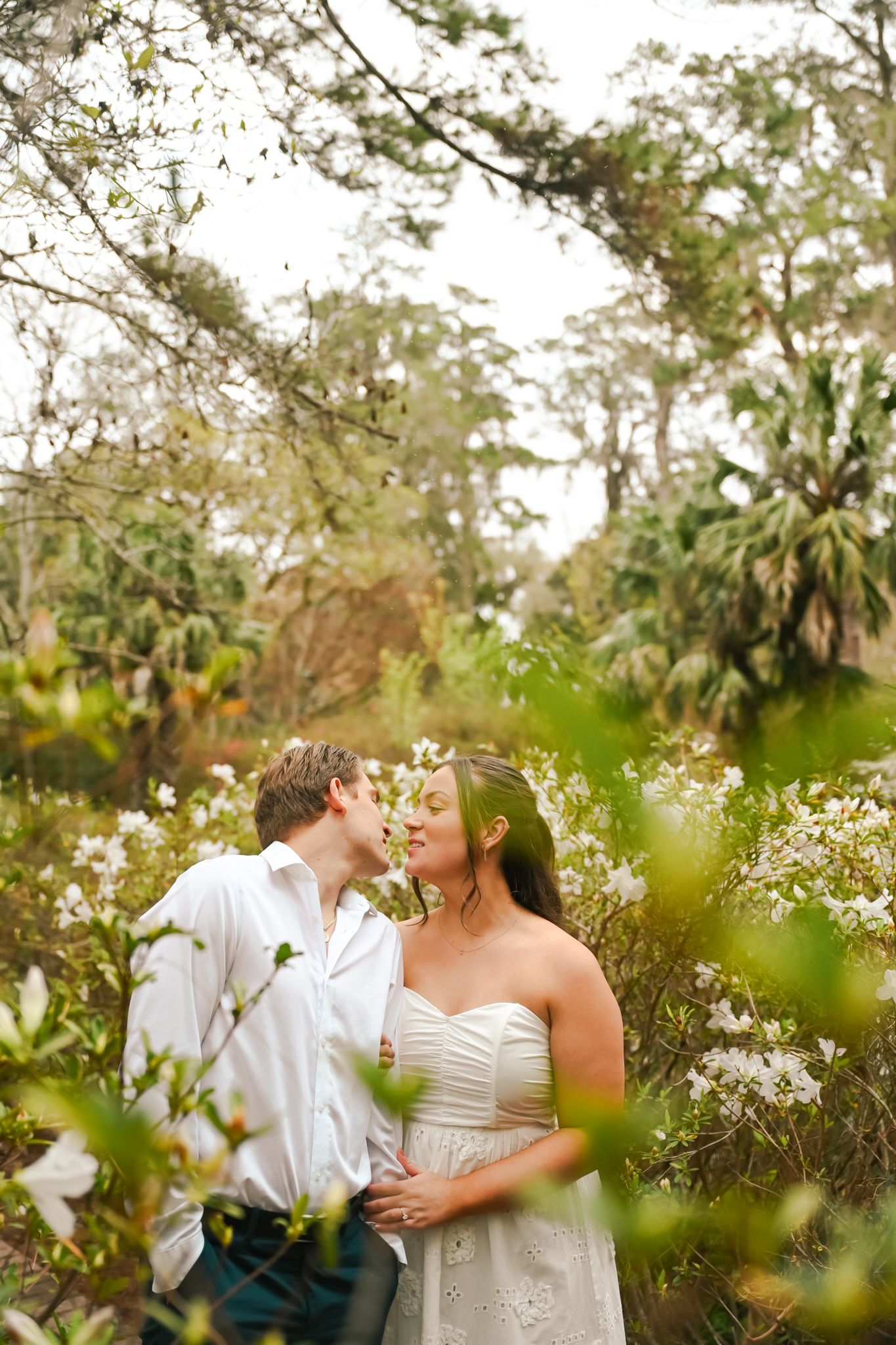 Romantic couple sharing a kiss among lush white flowering bushes in a dreamy outdoor garden setting.