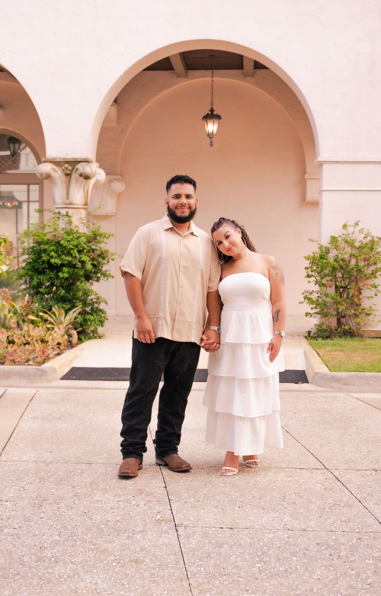 Couple posing together in front of arched building, man in beige shirt, woman in white tiered strapless dress.