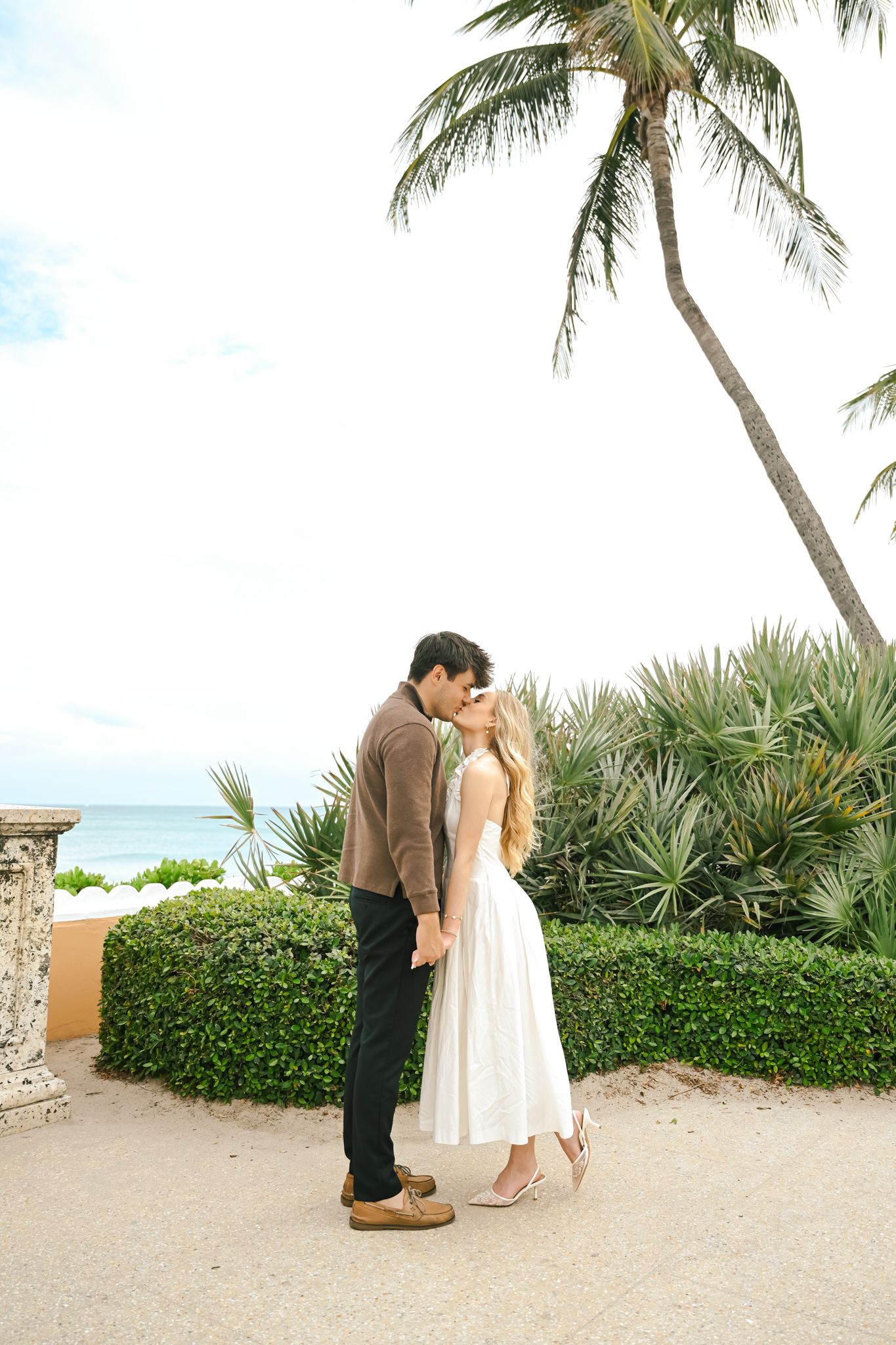 Couple sharing a romantic kiss near palm trees and ocean views in a tropical garden setting.