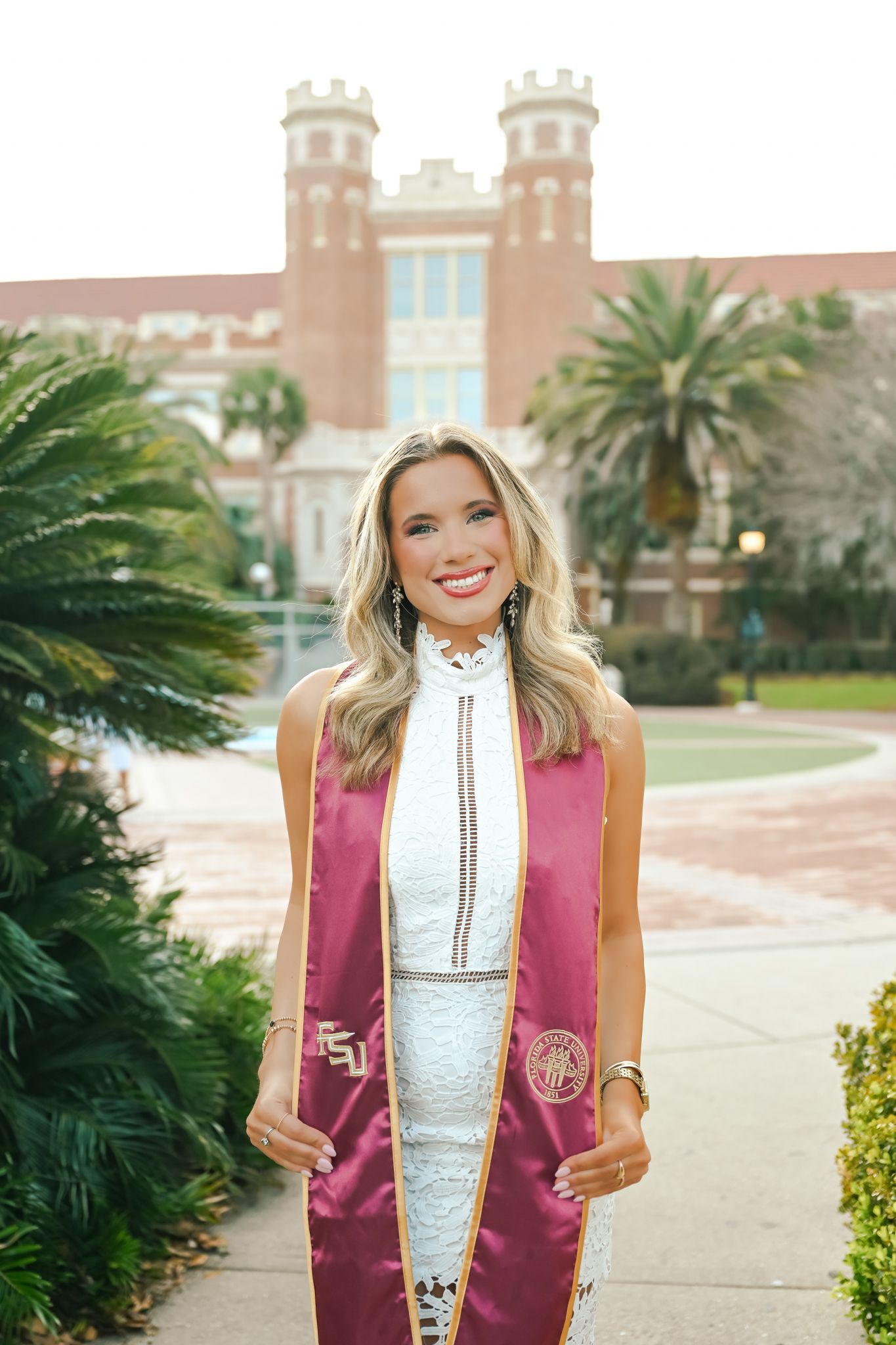 Graduate wearing burgundy graduation stole and white lace dress poses for photos on campus walkway.