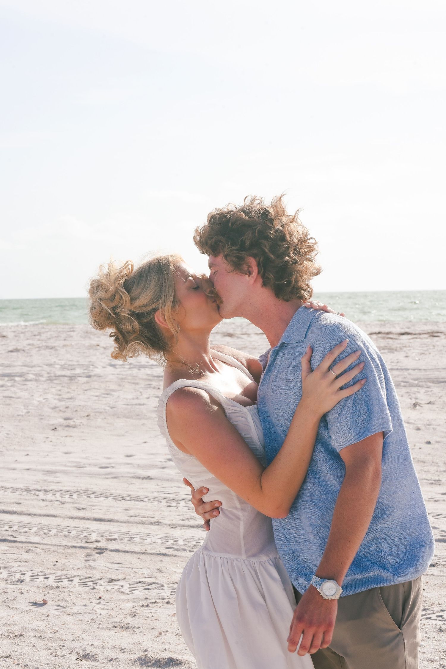 A couple shares a romantic kiss on a sunny beach, the woman in a white dress and man in a blue shirt.