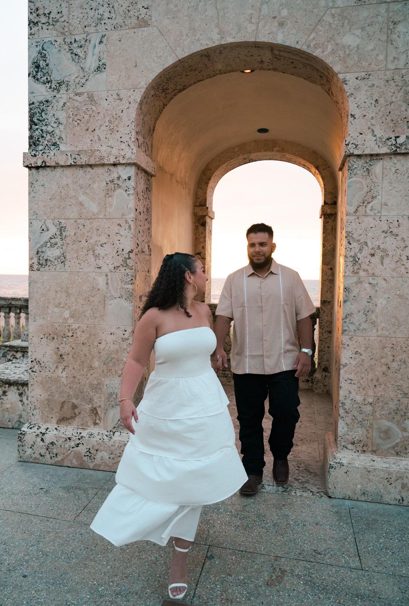 Bride in white strapless gown and groom in casual attire pose under stone archway at sunset venue.