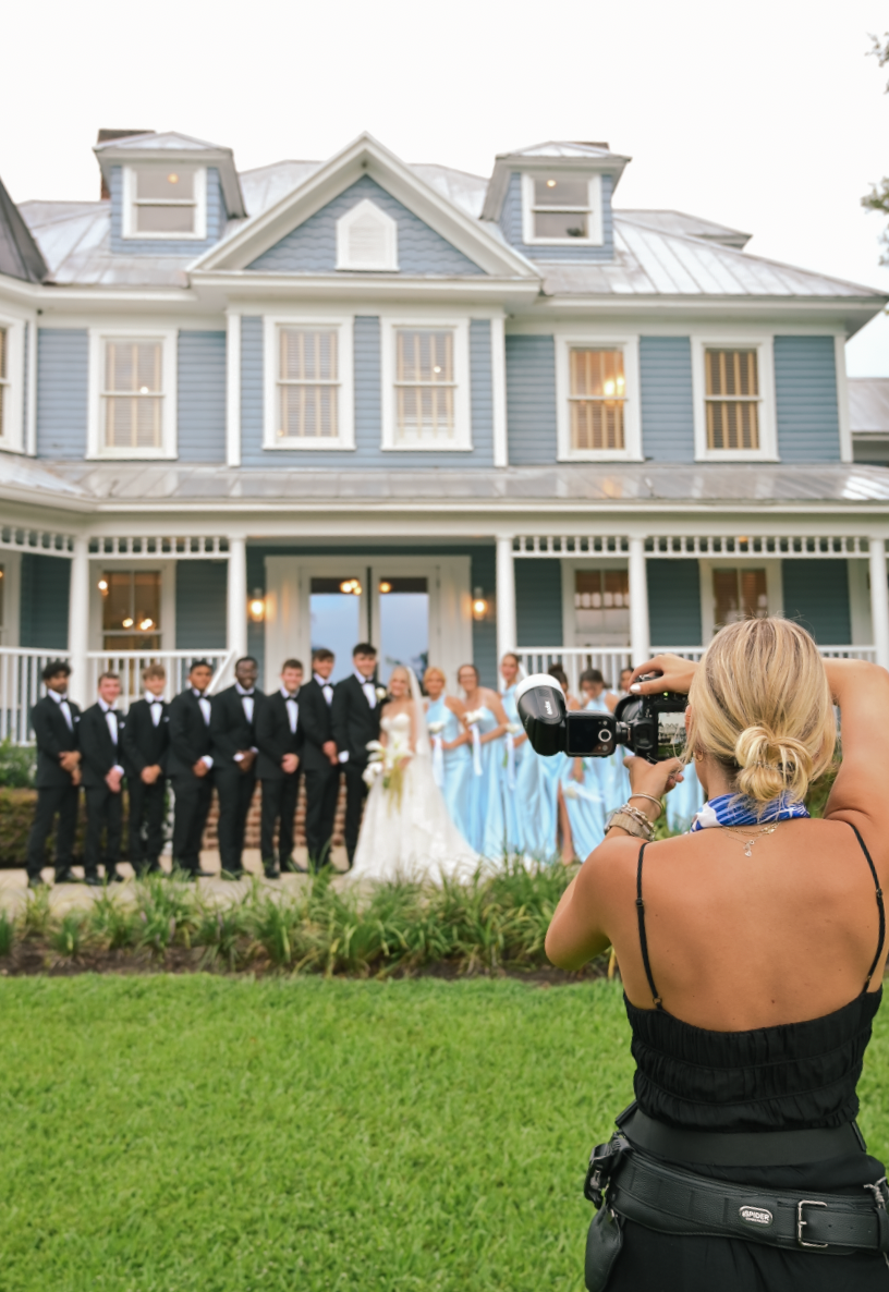 Female photographer capturing wedding party posed in front of elegant blue Victorian house with bridal party in light blue gowns.