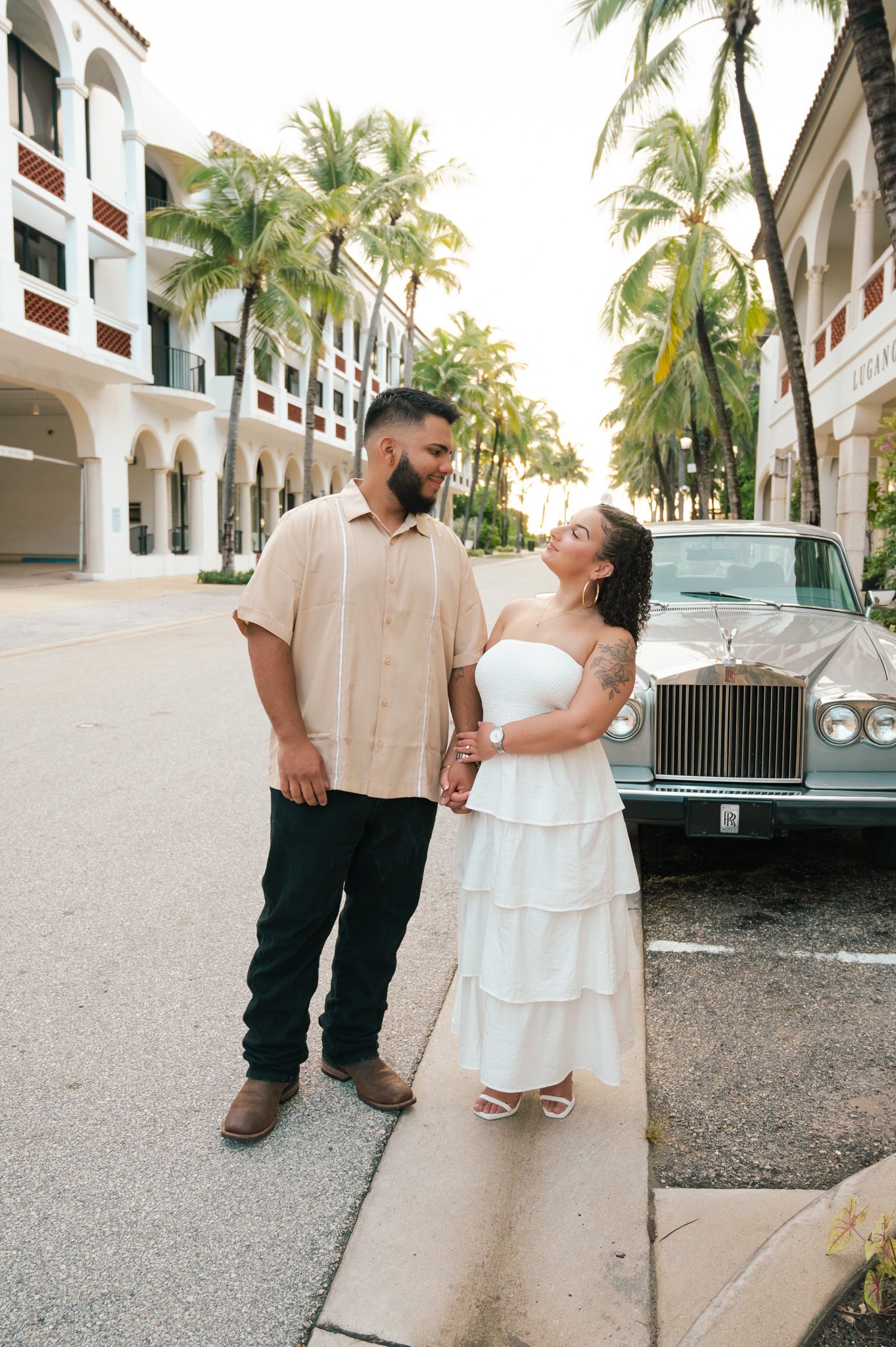 Couple poses casually by vintage car in front of palm-lined building.